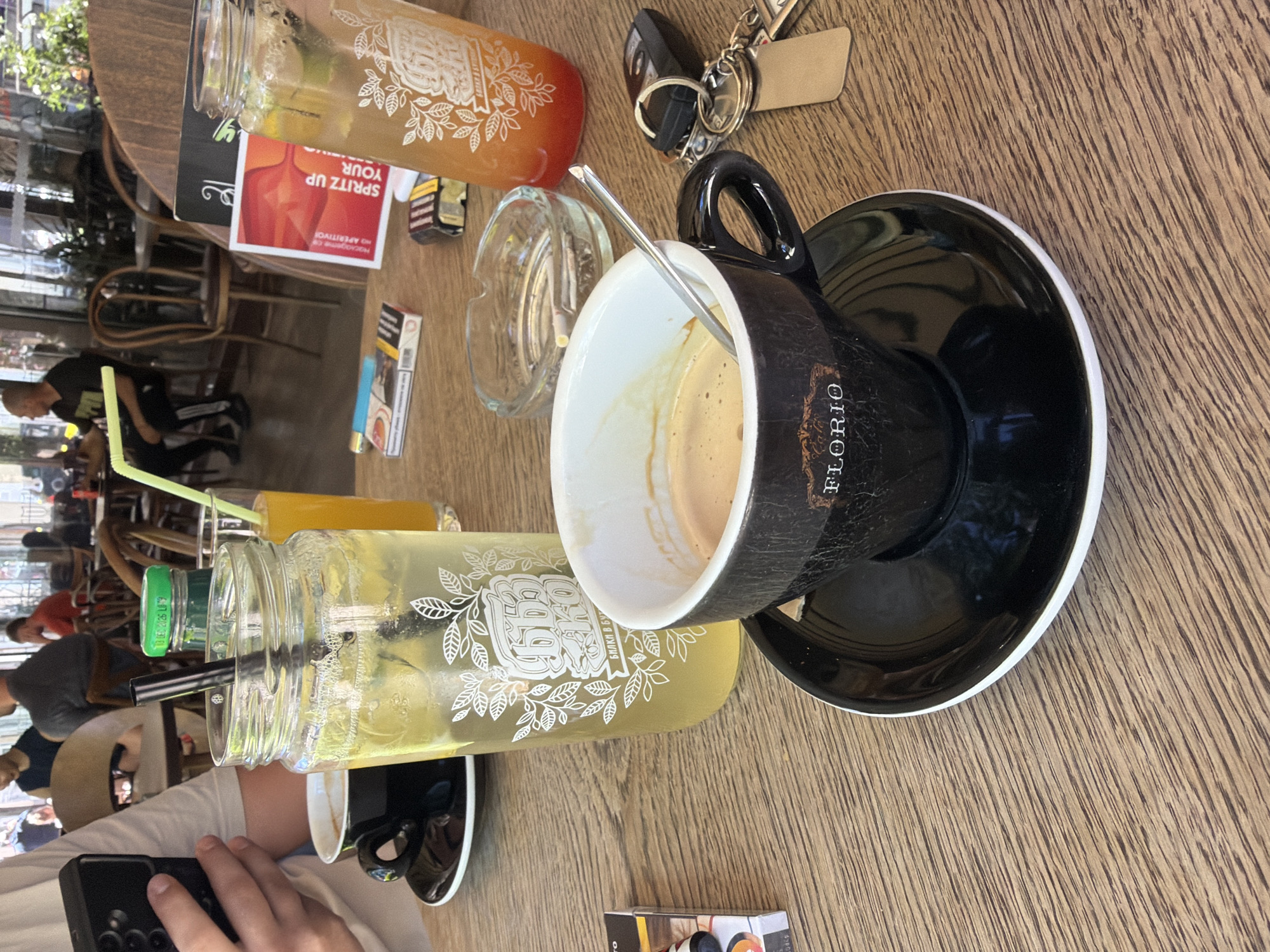 The scene captures a moment at Florio in Troyan, Bulgaria. A wooden table serves as the primary surface, displaying a partially consumed cup of coffee in a black mug with "FLORIO" inscribed on its side, resting on a black saucer with a white rim. A metal spoon is placed in the coffee cup. Alongside the coffee, there's a tall glass bottle filled with a light-colored beverage, possibly lemonade, and an orange-colored drink in a similar bottle, both featuring white leaf designs. A clear ashtray with cigarette butts, a pack of cigarettes, car keys, and a paper advertisement are also present. In the foreground, a person is holding a black smartphone, partially visible. The background reveals an outdoor café setting with blurred figures of people sitting at tables.