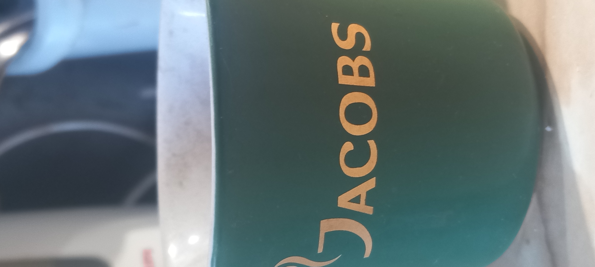 A close-up photograph shows a green coffee mug with the word "JACOBS" printed vertically in gold lettering. A stylized design, possibly representing coffee steam, is present below the first letter. Part of a white object, possibly the mug's handle or rim, is visible on the left. The mug is located near a kitchen countertop or surface that is lightly colored. A blurred stovetop with concentric circles is visible in the background on the left. The location is not discernible in this image and cannot be determined from this photograph.
