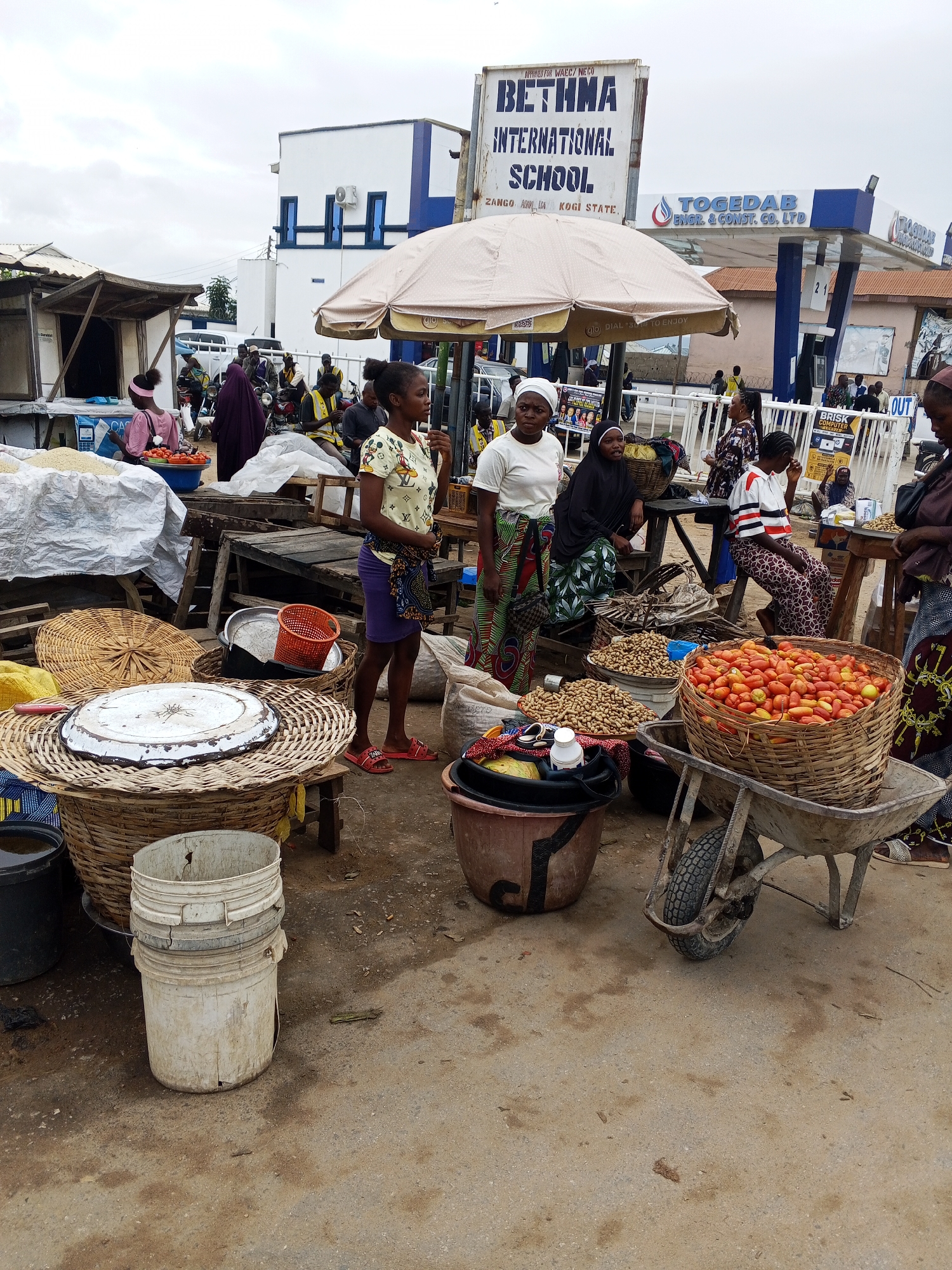 In Zango-ndaji, Nigeria, a market scene unfolds under an overcast sky, featuring a mix of vendors and customers. The foreground is dominated by market stalls laden with produce, primarily tomatoes and peanuts displayed in woven baskets. A wheelbarrow, partially visible, holds a large basket of tomatoes, suggesting recent transport or preparation for sale.
Several individuals are present, some actively engaged in selling or browsing, while others observe the surroundings. A woman in a yellow t-shirt and purple skirt stands near a stall, appearing to interact with a customer or attend to her wares. Another woman in a black hijab and patterned dress sits behind a wooden table, possibly a vendor. A young girl in a pink top and headband is seen in the background, near a stall selling what appears to be rice.
The market environment is characterized by a variety of materials, including woven baskets, plastic containers, and wooden structures. In the background, a building with signage for "BETHMA INTERNATIONAL SCHOOL" and "TOGEDAB ENGR. & CONST. CO. LTD" indicates the presence of local institutions. The overall impression is one of a bustling local marketplace, with everyday commerce taking place against an urban backdrop. The ground is a mix of dirt and concrete, typical of an outdoor market setting.