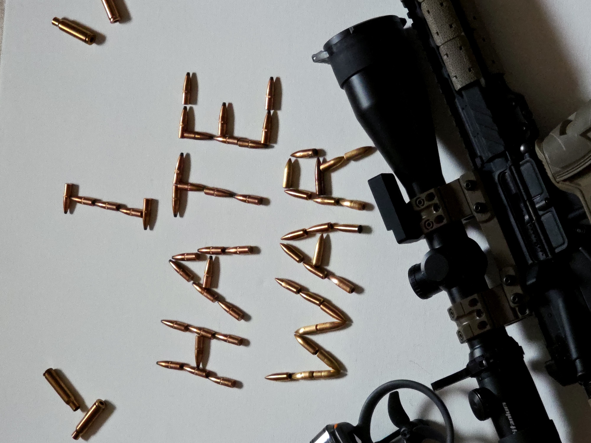 The image captures a firearm and ammunition arranged on a plain, light-colored surface. The central element is a phrase spelled out using numerous copper-colored bullet projectiles: "WE THE PEOPLE." The word "WE" is positioned on the upper left, "THE" in the upper center, and "PEOPLE" spans the lower central and right parts of the composition. Several complete cartridges, identifiable by their brass casings and copper tips, are scattered around the main text.
On the right side of the frame, a significant portion of a modern sporting or tactical rifle is visible. The rifle is primarily black with accents of tan or flat dark earth (FDE) on its handguard and possibly other furniture components. A large, black scope is mounted on the rifle, with the word "Titanium" clearly visible on its side. Below the rifle, a small, black circular object with propeller-like blades is partially seen. The scene is set indoors, under ambient lighting, without any specific time of day or external location cues. No individuals are present in the image.
