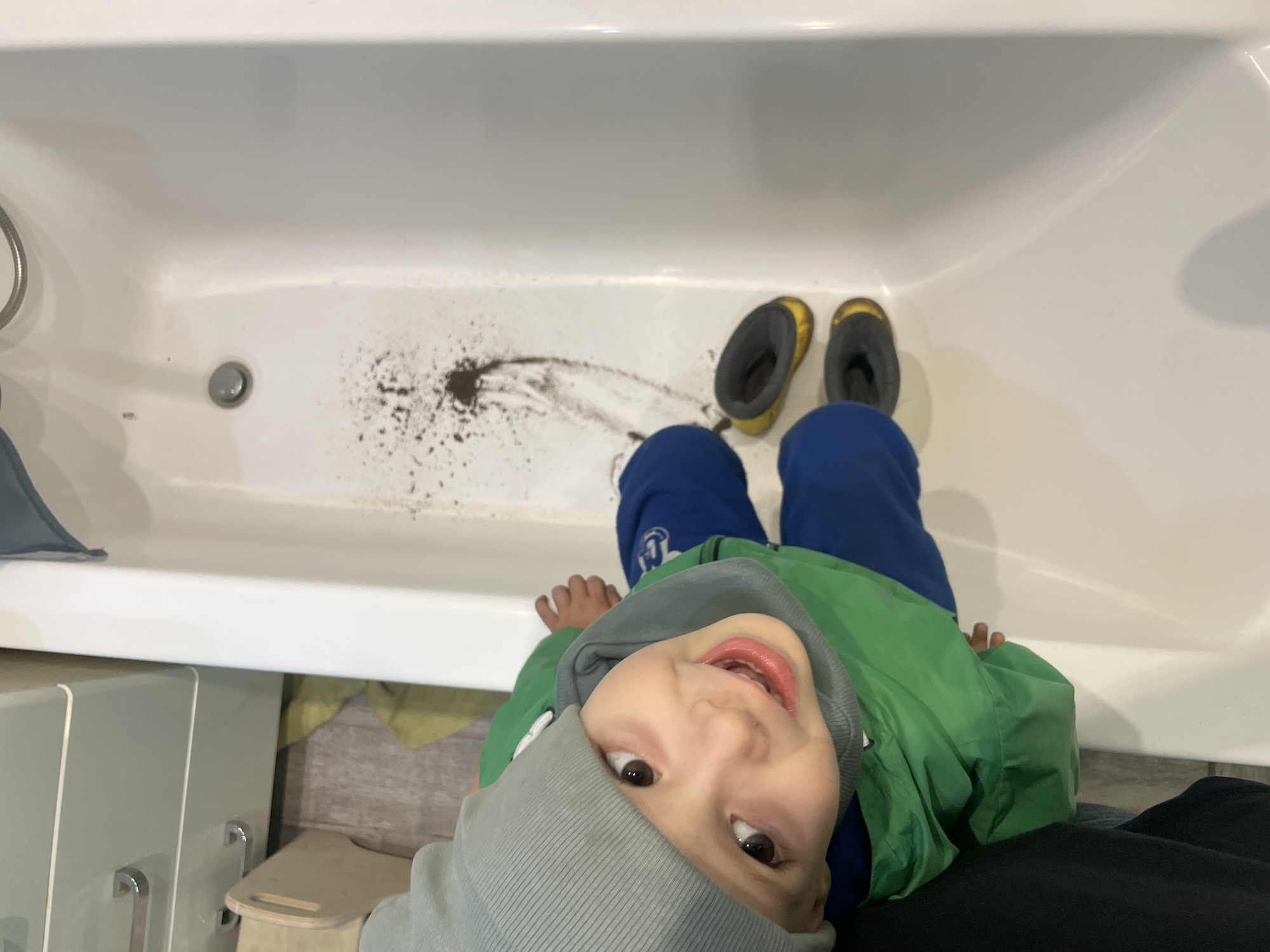 A child wearing a gray hat, green jacket, and blue pants is sitting in a white bathtub. The child is looking up at the camera and smiling. There are brown clumps in the bathtub near the drain and a pair of yellow-and-gray rain boots are near the child's feet. A gray cabinet is visible next to the bathtub. The location is Unknown, Unknown.