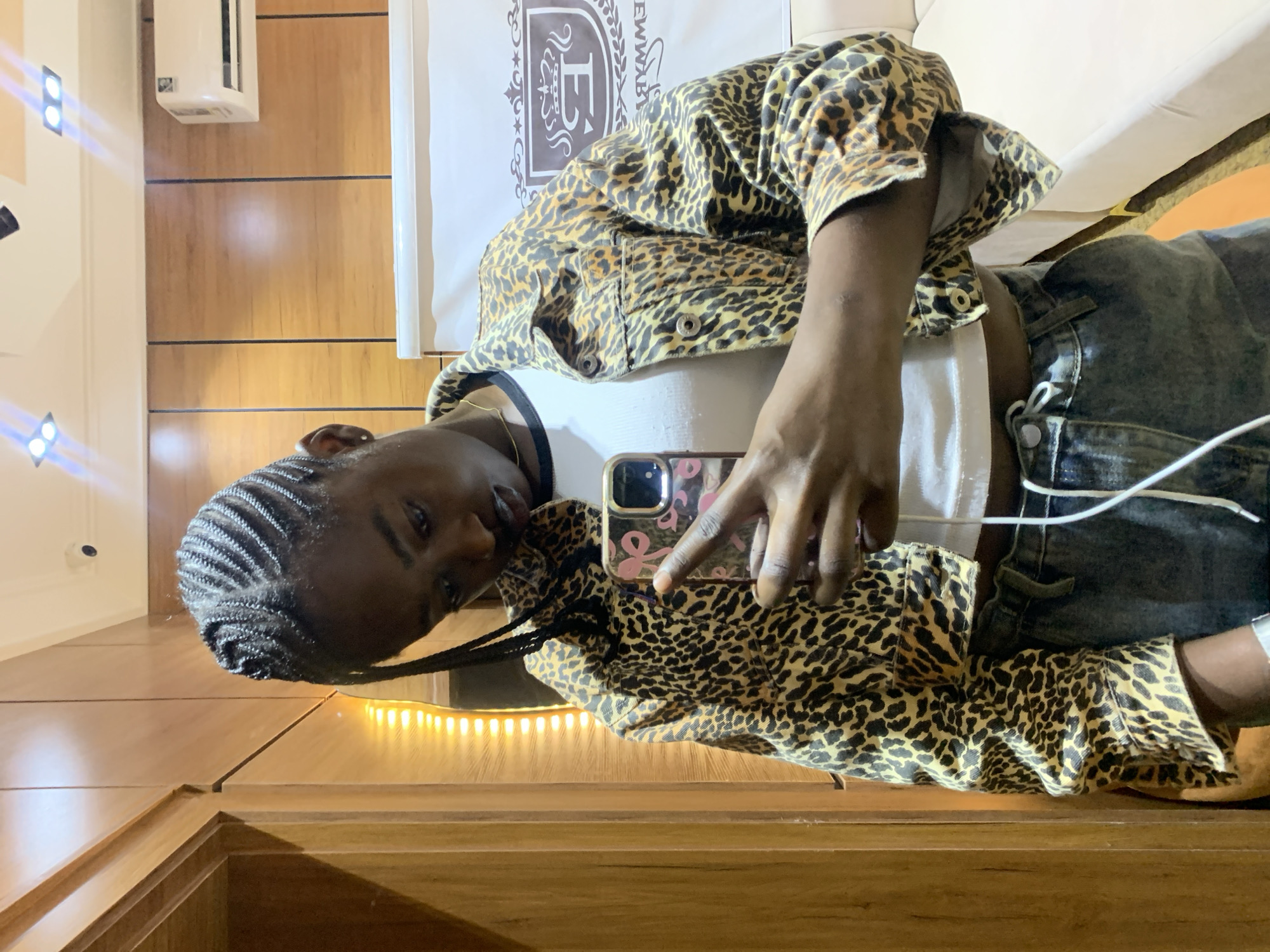A medium shot shows a young Black woman with braided hair taking a selfie in a room that appears to be a hotel or guest room. She is wearing a leopard-print cropped jacket over a white tank top and denim jeans. She is holding a smartphone in her right hand, with her left hand resting on her chest. The background includes wooden paneling on the walls, a large white air conditioning unit, a white screen or banner with a crest logo, and part of a light-colored sofa. The lighting suggests an indoor setting, possibly during the evening. No specific location in Gura Top, Nigeria, is identifiable from the visual cues in the image.