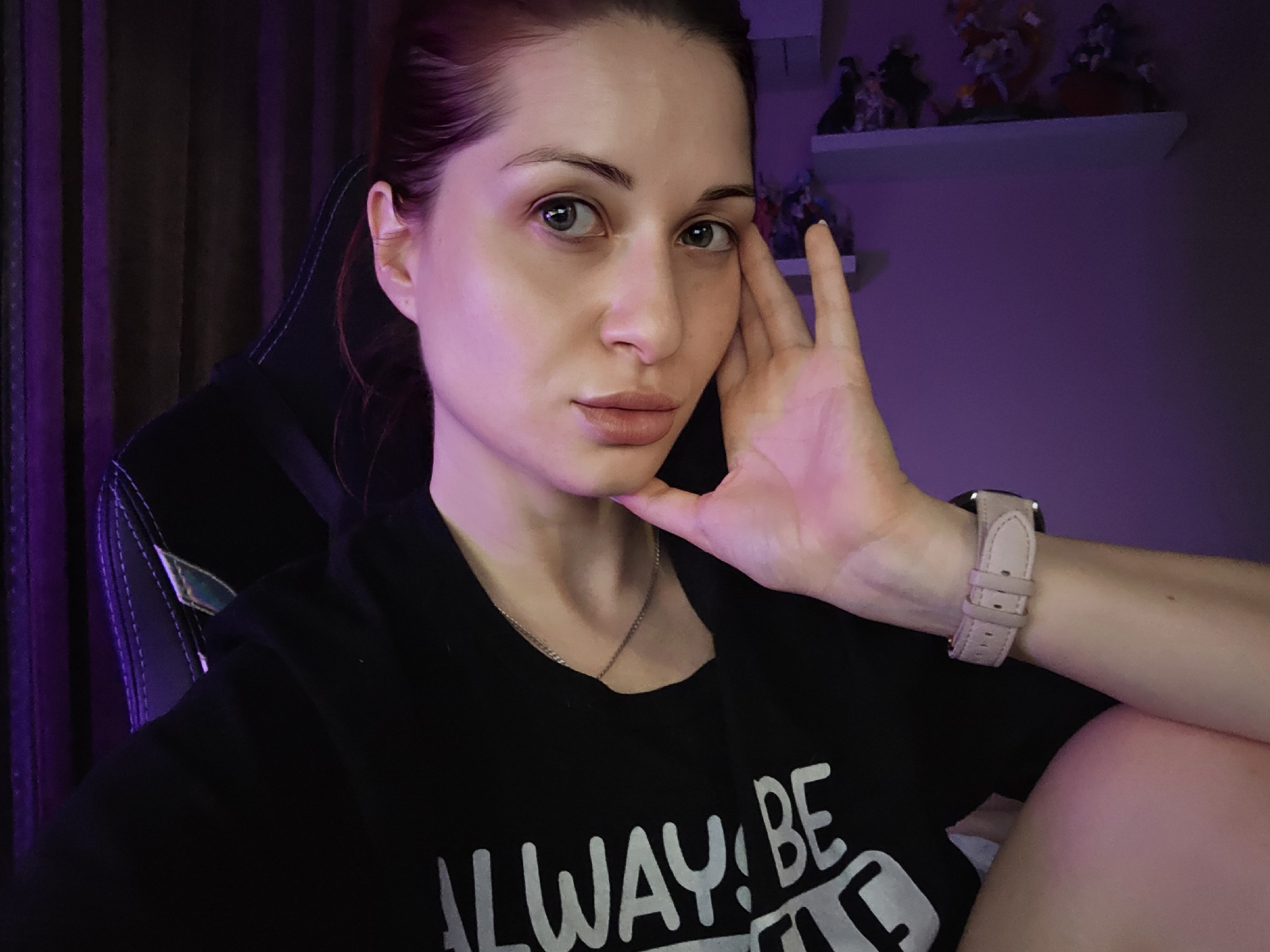 A woman with red hair pulled back poses for a selfie. She rests her left hand against her face. She has fair skin, light eyes, and is wearing a silver chain necklace and a light pink wrist watch. She is wearing a black t-shirt. She is sitting in a black and grey gaming chair. The background includes dark curtains on the left, a wall lit with purple light, and a shelf with figurines. There is no evidence to indicate the location is Burgas, Bulgaria.