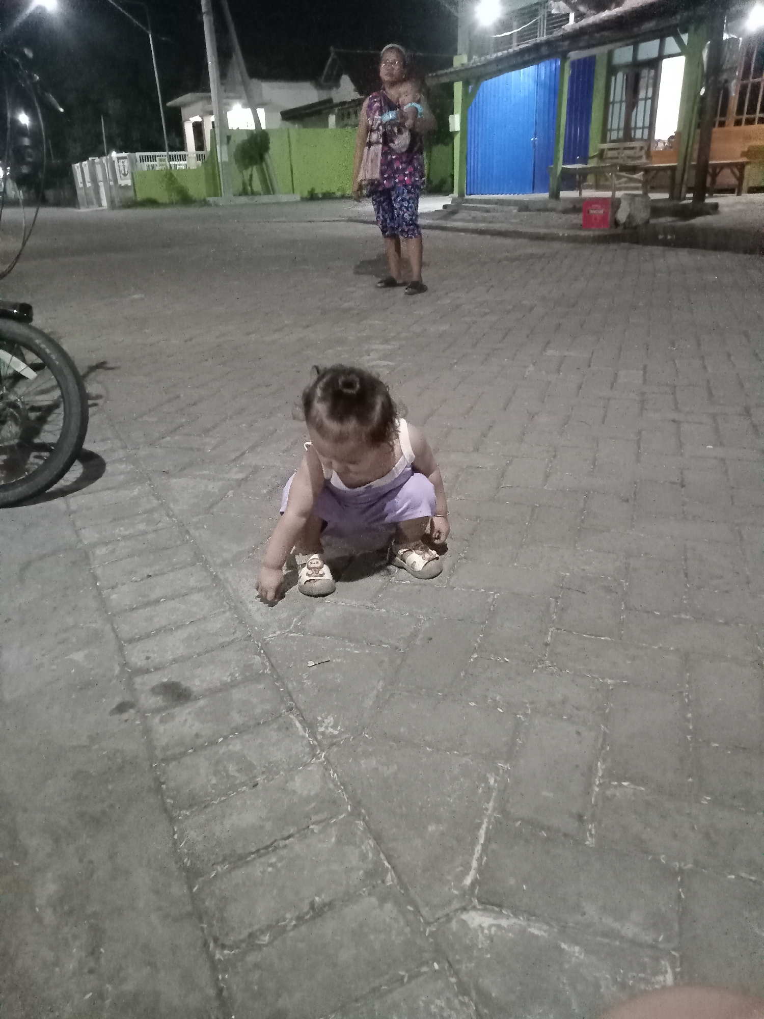 A young child in a light purple outfit is crouching on a paved street surface in Bungur, Indonesia. The child appears to be looking at something on the ground. In the background, an adult woman is standing, possibly holding a baby or child in her arms. A bicycle wheel is visible on the left side of the image. The scene is set in a residential area, with houses and streetlights in the background. One house features green walls and a blue door or shutters. The ground is made of light grey rectangular pavers. It seems to be dusk or nighttime based on the light conditions.