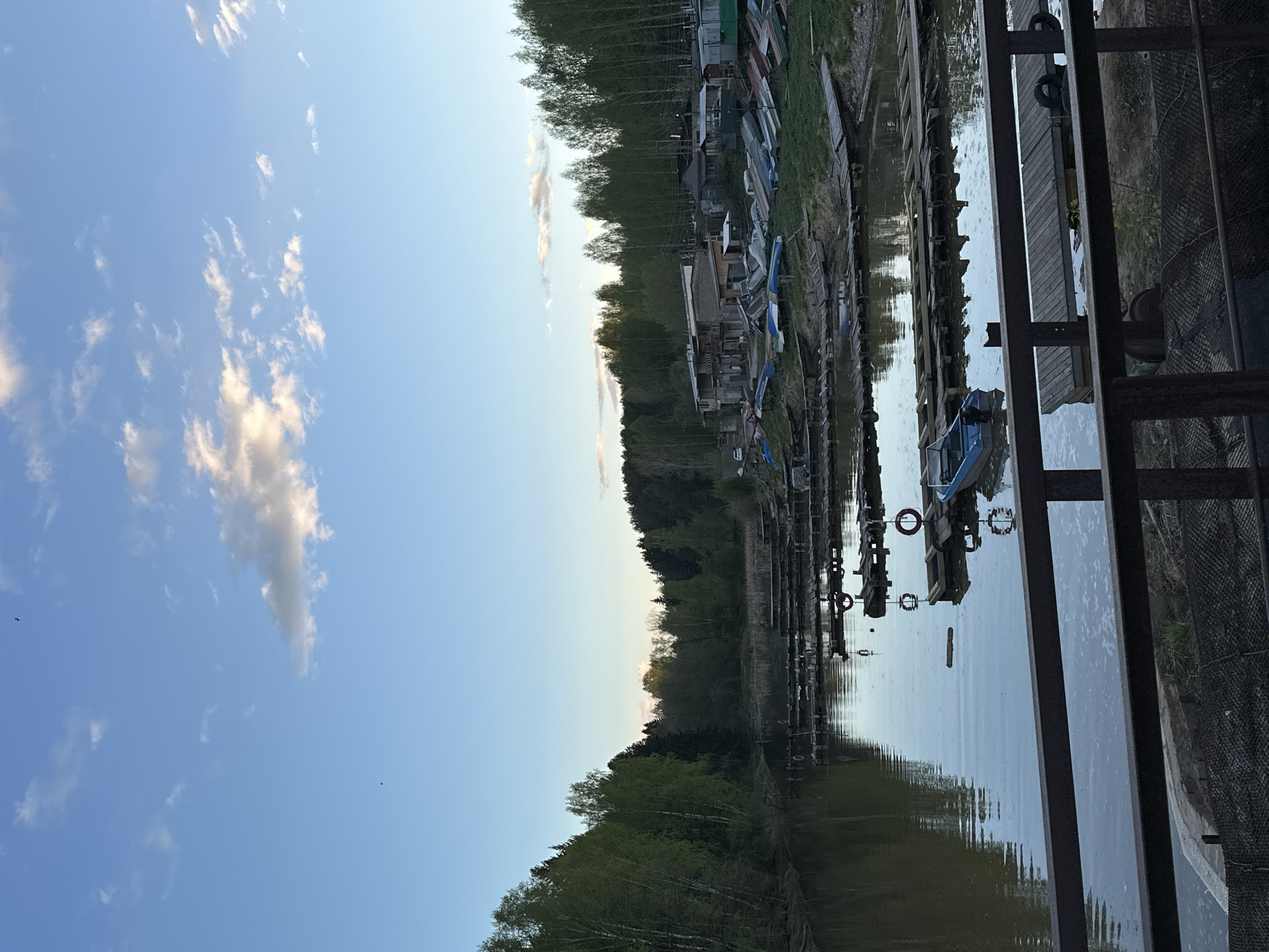 The image depicts a view of a waterfront area in Chaikovsky, Russia. The scene is oriented with the sky to the left and water on the right. Above, there's a blue sky with scattered white clouds.
Along the top of the image, and reflected in the water, is a line of trees and what appears to be a cluster of small buildings or sheds. Various small boats are moored alongside the water.
In the bottom right foreground, there are metal structures, possibly part of a dock or pier.