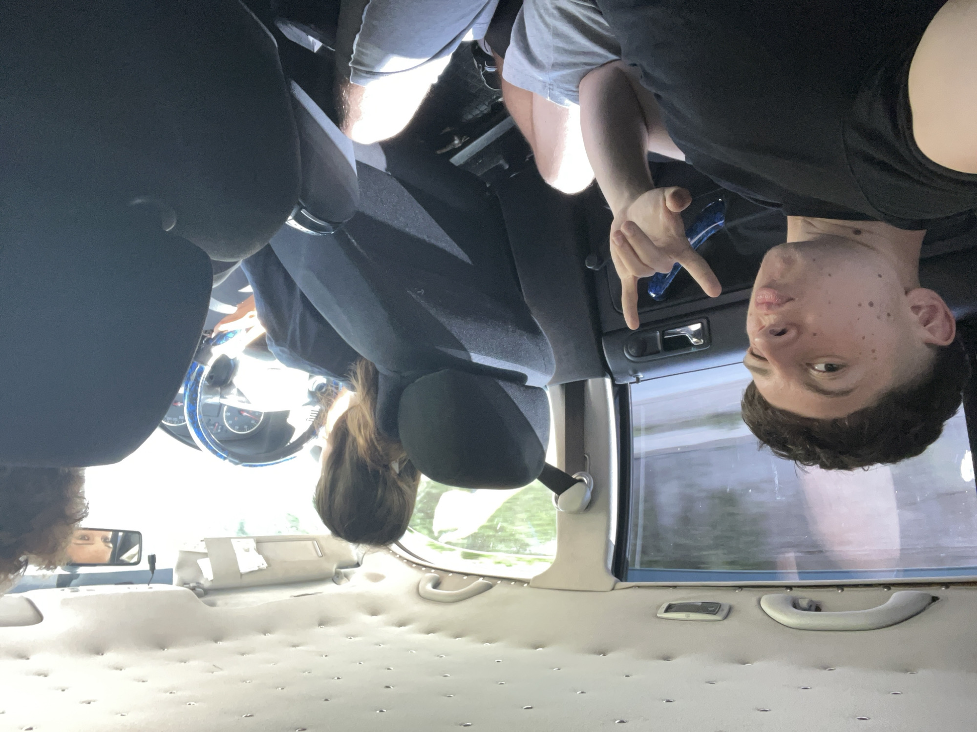 The image shows the interior of a vehicle, likely a car, with four people inside. The viewpoint is from the back seat, looking towards the front. The image is rotated 90 degrees clockwise, making the people appear upside down.
In the foreground, the plush, tufted interior of the back seats is visible. A person's head with curly brown hair is visible in the lower left corner, facing towards a rear-view mirror. The mirror reflects a person's eyes.
In the middle ground, the back of the driver's seat and part of the passenger seat are visible. A person with long brown hair is seated in the back, facing forward. A person in the passenger seat is wearing a grey t-shirt and shorts.
In the background, the front of the car is visible. The driver, a young man with short brown hair and freckles, is in the driver's seat. He is wearing a black t-shirt and is making a "rock on" hand gesture. He is looking directly at the camera with a playful expression. The steering wheel and dashboard of the car are visible. The window shows a blurry view of trees and greenery, suggesting an outdoor setting, possibly a road trip. The lighting suggests it is daytime, likely a sunny day, given the bright light illuminating the interior.
There are no visible text elements in the image. The overall mood appears to be casual and friendly, with the subjects seemingly enjoying their time together. The location is Vlahovo, Bulgaria.