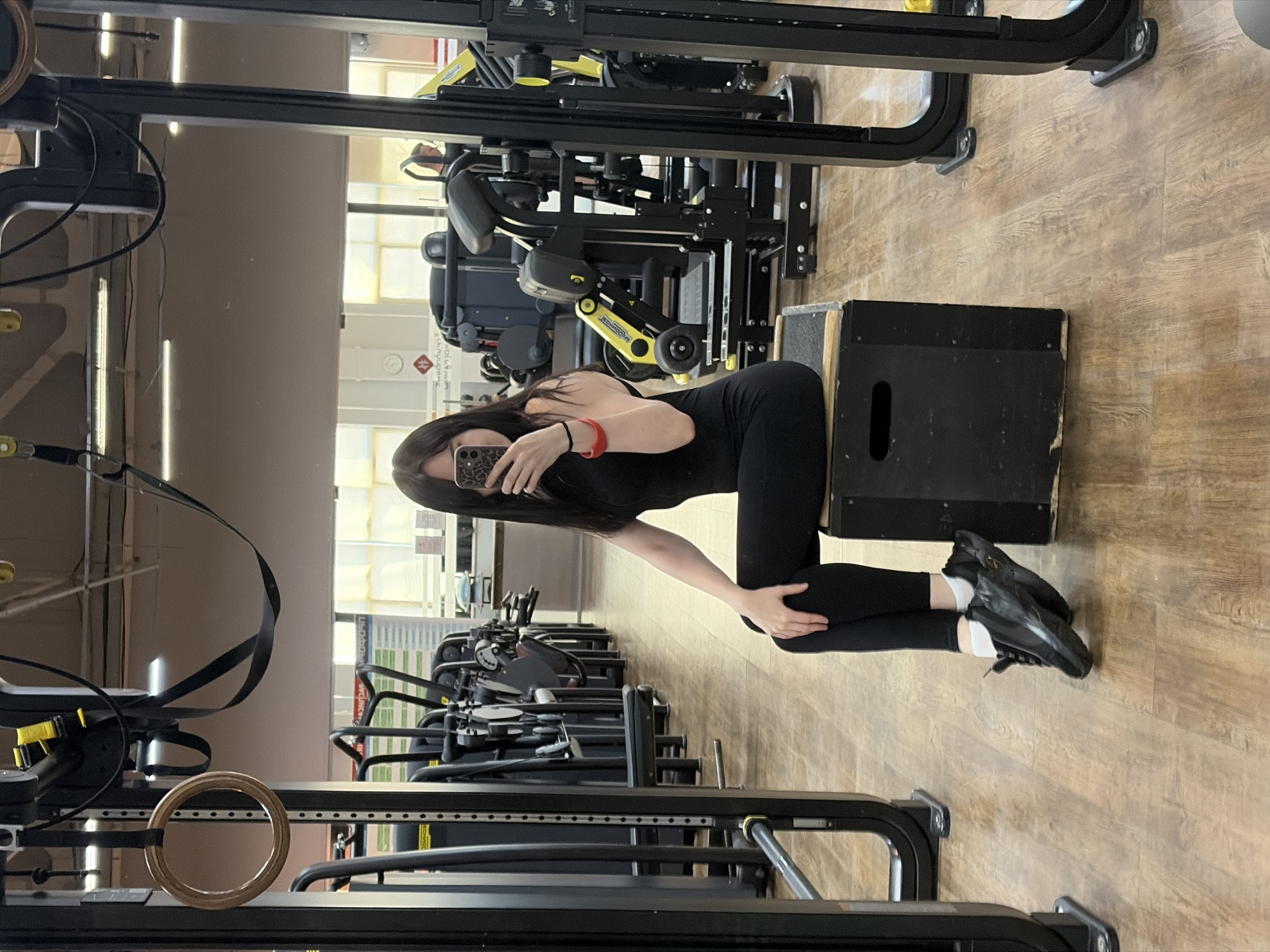 A woman in black athletic wear is kneeling next to a black plyometric box inside a gymnasium. She is holding a phone in her left hand as if taking a selfie. The background shows various exercise machines arranged in rows. The location is likely a fitness center in Chaikovsky, Russia.