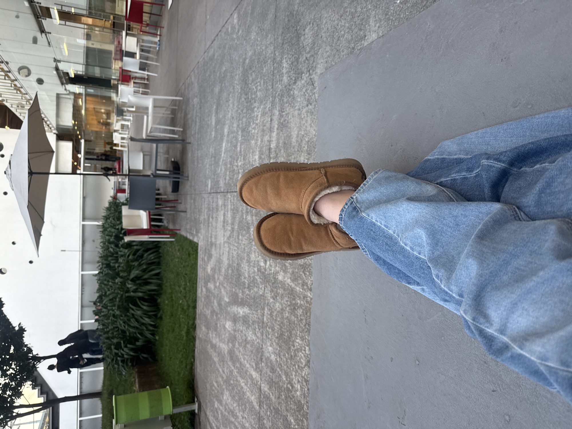 The image displays a first-person perspective view of a person's lower legs and feet in an outdoor public plaza, identified as a public area at the base of the Torre Virreyes complex (Pedregal 24, Molino del Rey, Miguel Hidalgo, 11040 Mexico City, CDMX, Mexico).
In the foreground, the observer's legs are extended and crossed at the ankles, resting on a flat, dark gray concrete or stone surface. The person wears light blue, loose-fitting denim jeans and tan, fleece-lined, ankle-height boots.
Beyond the immediate foreground, a light gray, textured concrete or paved surface extends into the midground. To the left, a patch of green grass with dense, dark green foliage (possibly liriope) is visible. A light green, perforated metal waste bin on a pole stands at the edge of this grassy area. Further back on a raised grassy mound, against a modern white building wall, several silhouetted figures (at least three individuals) are seen walking or standing near a tree and a light-colored outdoor staircase.
To the right and center midground, an outdoor dining or seating area is arranged. It features numerous tables with dark gray bases and white tops, accompanied by a mix of white, gray, and red outdoor chairs. One large, light gray outdoor umbrella with a central pole stands over a table in this area.
The background consists of a modern, multi-story building characterized by extensive glass facades that reflect ambient light and interior elements, indicating a commercial or office structure. The overall scene suggests a late afternoon or early evening setting due to the soft lighting and the partial silhouetting of figures.