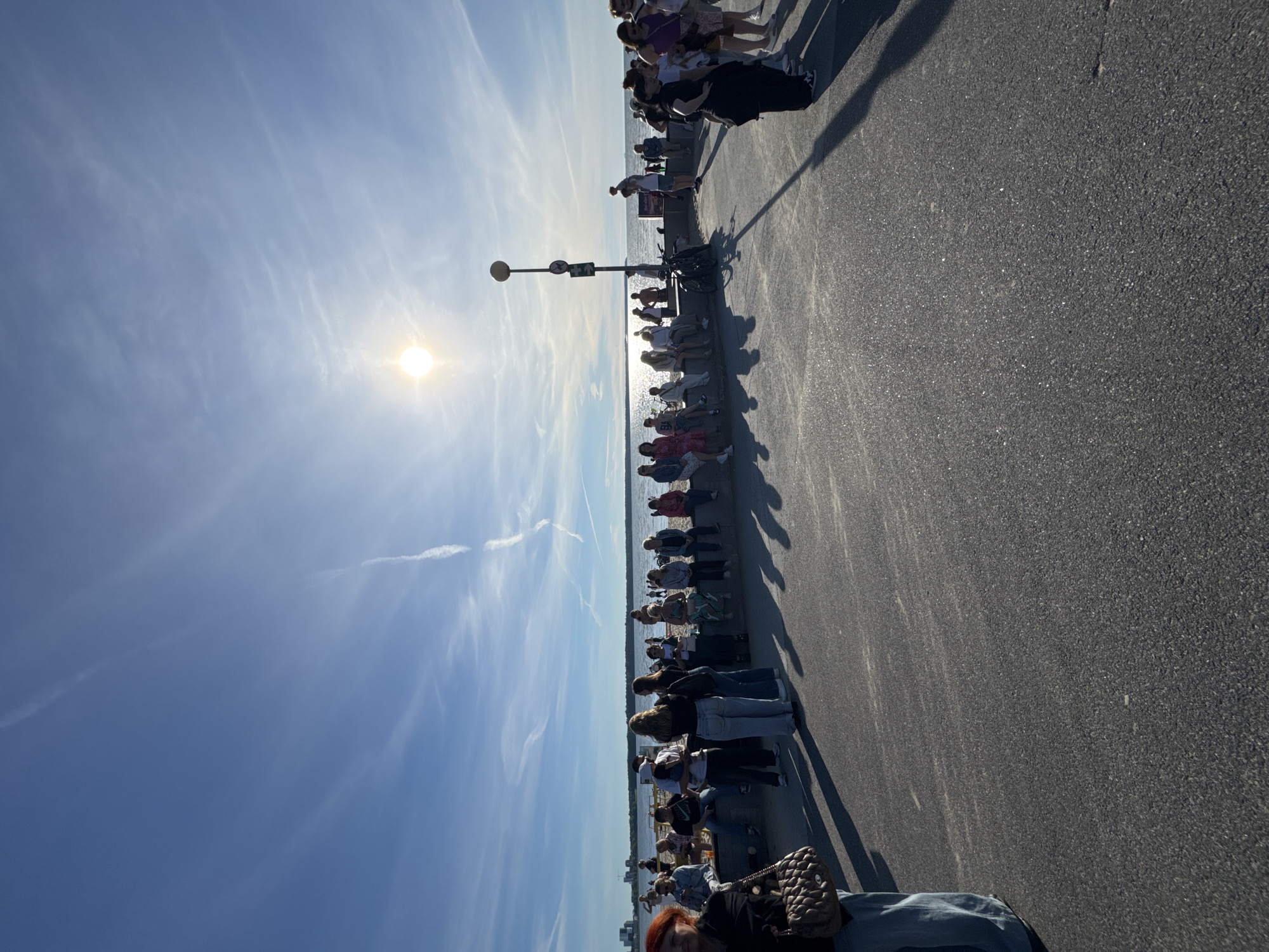 The media file depicts a sunny day with a clear blue sky and wispy clouds, suggesting it is daytime, likely late morning or early afternoon, given the angle of the sun and the shadows. The scene is an outdoor promenade next to a body of water, possibly the Baltic Sea, with a distant shoreline visible. A line of people is gathered along the promenade, facing the water.
The subjects are primarily people, dressed in casual summer attire. They appear to be standing or sitting in a relaxed manner, some engaging in conversation, while others are looking out at the water. The activity seems to be a casual gathering or observation of the scenery. There are a few bicycles parked along the promenade, indicating it is a public area accessible by bike. A lamppost with a circular lamp and a sign are also visible.
Notable details include the bright sunlight casting long shadows, highlighting the texture of the asphalt promenade. The water appears calm, reflecting the sky. The overall mood seems peaceful and leisurely.
There is no visible text within the image. The location context provided is Tallinn, Estonia, which aligns with the possibility of a seaside promenade.