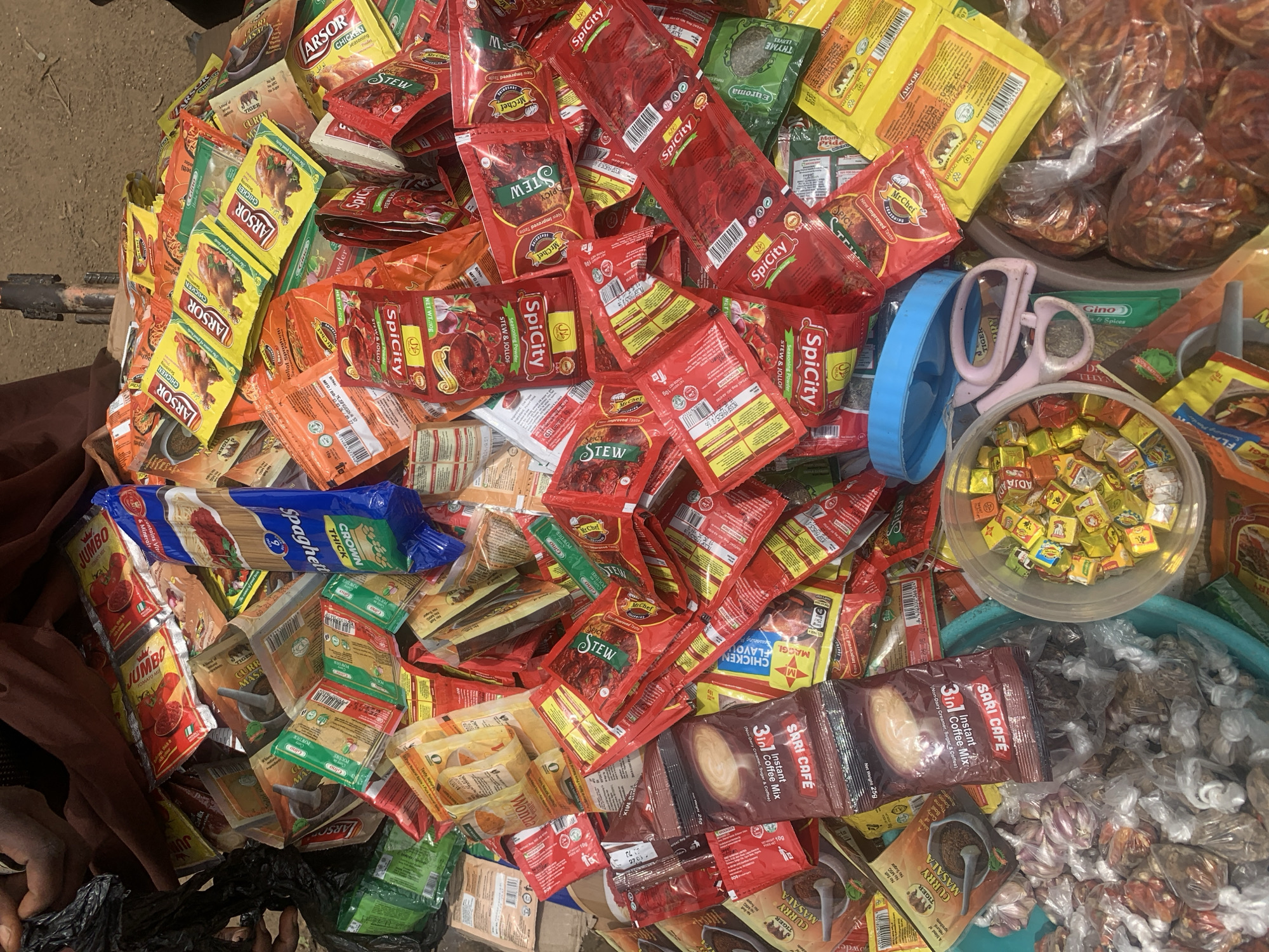 The image shows a street-level view of a vendor's collection of seasonings and food items, likely for sale in Jos, Nigeria. A variety of packaged seasonings are visible, including numerous red packets labeled "SpiCity Stew & Jollof," yellow packets of "LARSO" chicken seasoning, "Mr. Chef" seasoning, packets of "Tiger Curry Masala," packets of "3in1 Sari Cafe Instant Coffee Mix," as well as "JUMBO Tomato Mix" and "MAGGI Chicken Flavoured." A blue package of "Crown Thick Spaghetti" is also visible. Beside the packets is a clear plastic container filled with small, bouillon cubes. A pair of scissors with light pink handles and a blue measuring cup are resting among the items. To the side of the pile are small, clear plastic bags filled with various dried spices and ingredients. A dark-skinned person is partially visible on the left side of the frame. The overall environment suggests an outdoor market setting with a slightly dusty surface visible.