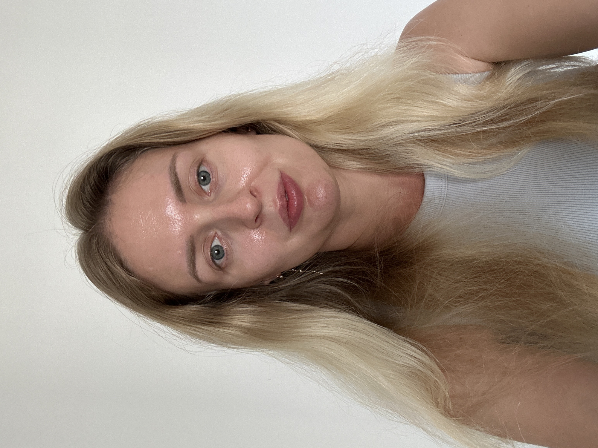 A frontal headshot and upper-body portrait features a single individual, an adult female. She has long, blonde hair, parted down the middle, which falls over her shoulders. Her eyes are blue, and her skin is light-toned, showing some natural sheen on her forehead, nose, and chin. She has full lips with a subtle pink tint and wears a small, thin, gold-colored dangling earring on her left ear. Her expression is neutral with a slight hint of a smile. She is dressed in a light grey ribbed tank top. The background is a plain, light-colored (white or off-white) wall, devoid of any discernible features or objects. The lighting is even and appears to be frontal. There is no information within the image to determine a specific geographical location such as the city of Petah Tikva, Israel.