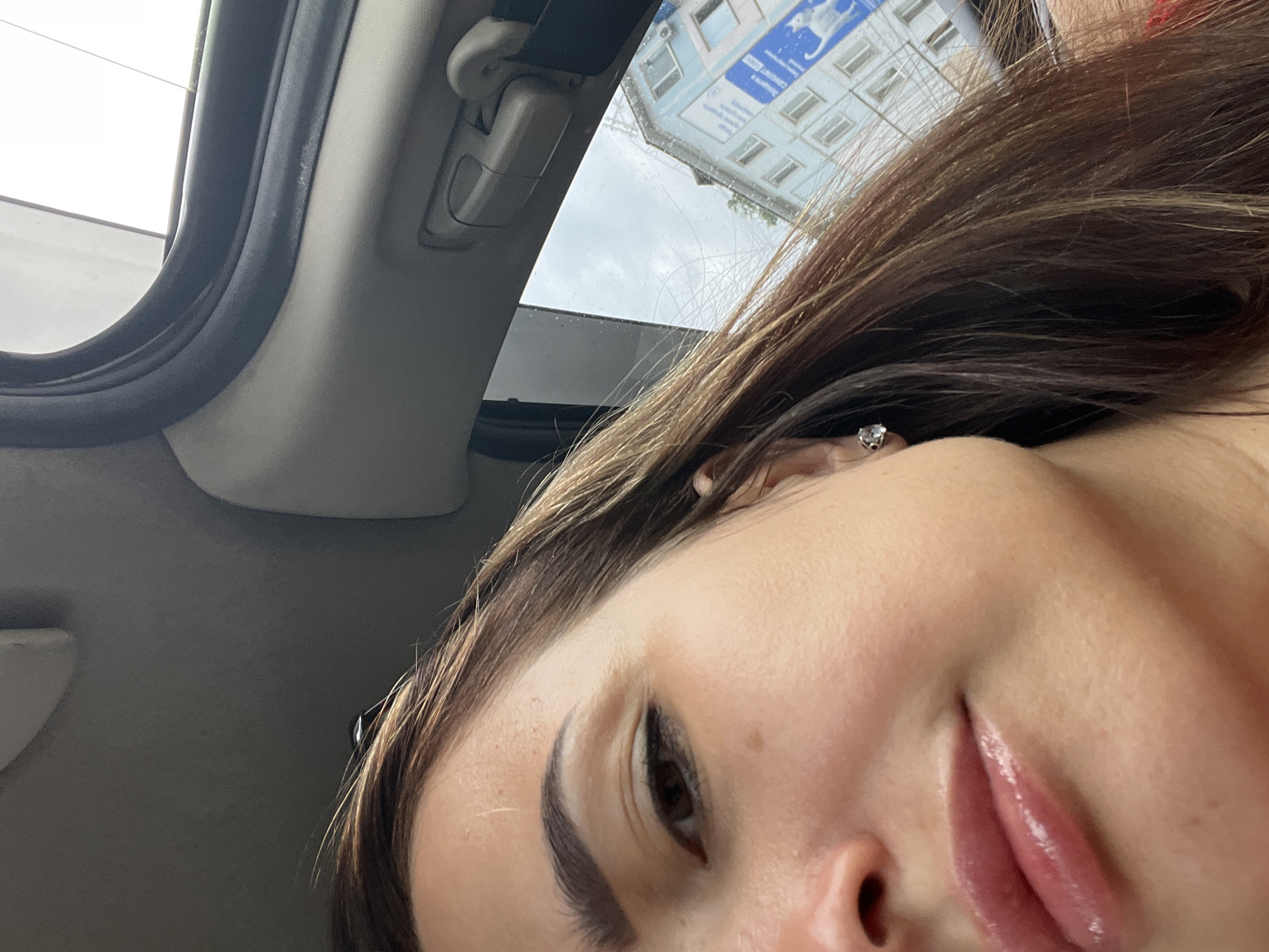 The media file is a close-up, overhead shot of a young woman's face, taken from inside a vehicle. Her head is tilted, and she is looking towards the camera. The sunroof of the car is open, revealing a cloudy sky and the top of a building with multiple windows. The building appears to be an apartment complex or office building. The woman has long, brown hair with lighter highlights, and she is wearing a diamond stud earring. Her lips are slightly parted and appear to be glossy. There is some visible text on the building in the background, but it is not clearly legible. The overall impression is that of a candid, casual snapshot taken during the daytime.