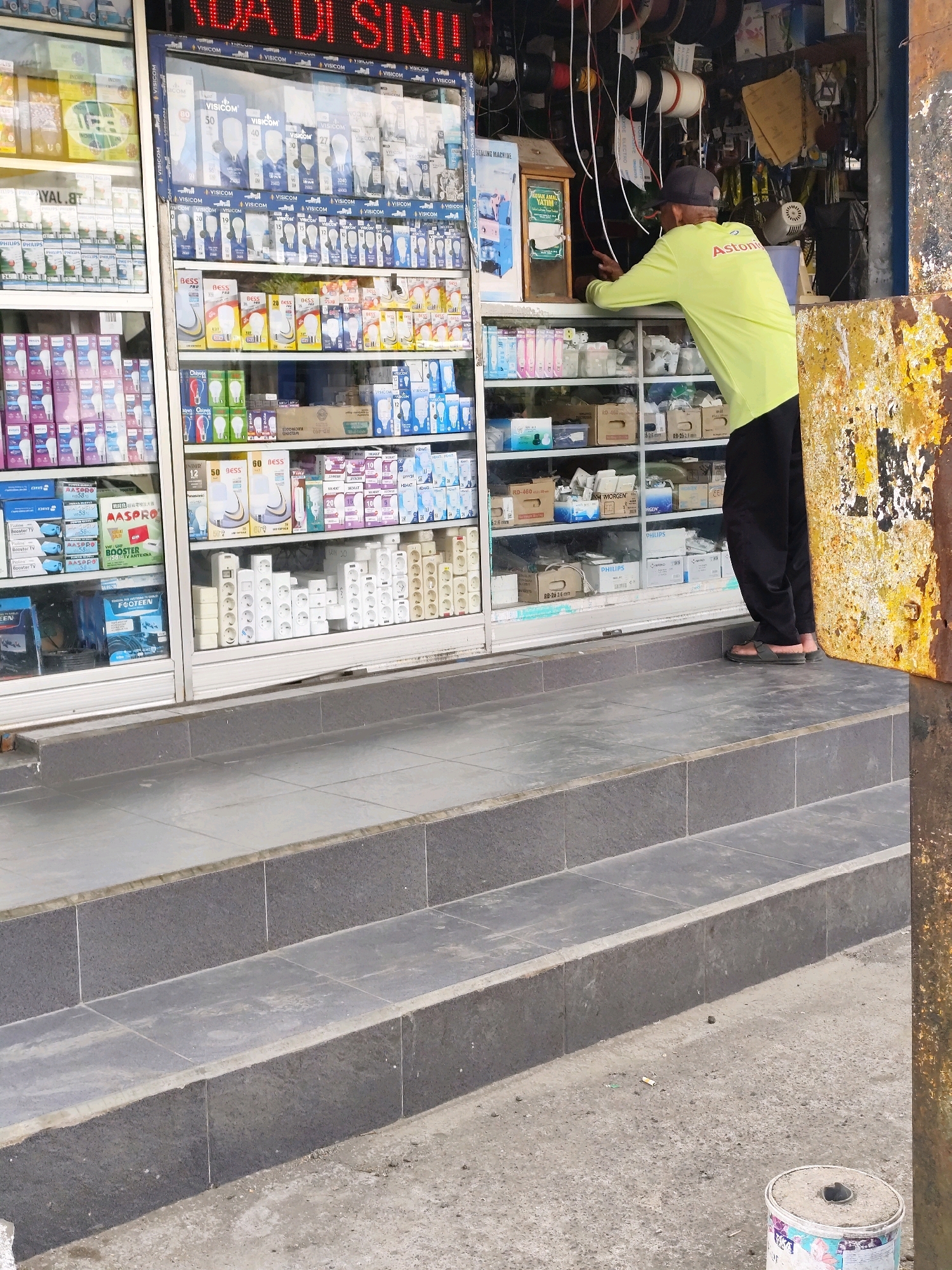 The image shows a street-level view of an outdoor shop in Jambewangi, Indonesia, featuring a display of electrical goods and supplies. A man wearing a neon yellow long-sleeved shirt and black pants, with a dark baseball cap, is standing with his back to the camera, leaning over a shelf inside the shop. He appears to be interacting with the merchandise. The shop's facade includes glass display cases filled with various electrical items, including light bulbs, power strips, and packaging for electronic components. The items are arranged on shelves that extend from the floor upwards. The shop is situated on a street with a concrete sidewalk and steps leading up to the shop entrance. The overall environment suggests a commercial area in a tropical climate, with evidence of wear and tear on the building's exterior, including rusted metal elements on the right side of the frame. The ground in front of the shop is paved with concrete and scattered with debris, including a discarded cigarette butt and a paint can. The lighting indicates it is daytime.