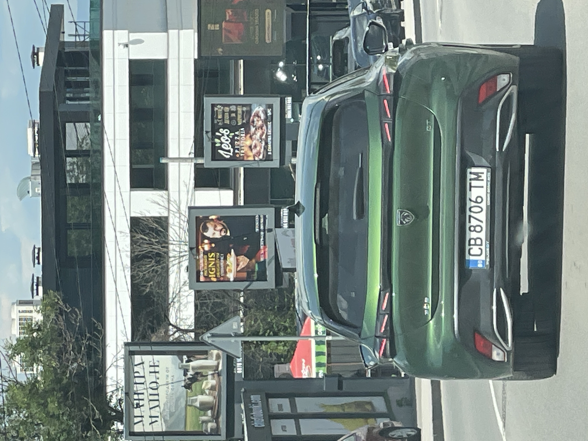 The image shows the rear of a green Peugeot 308, license plate CB 8706 TM, driving on a city street in Sofia, Bulgaria. Several billboards are visible in the background. One billboard, to the left, advertises dairy products with the text "МАНТРА МАЛОЛЕ". Another billboard, further right, advertises "Leo's PIZZA TRATTORIA" with an image of pizza. To the left of the Leo's billboard, another billboard displays an image of a man holding a plate of food, with text that includes "IGNIS". The environment appears to be a sunny day in an urban setting, with buildings and trees visible.