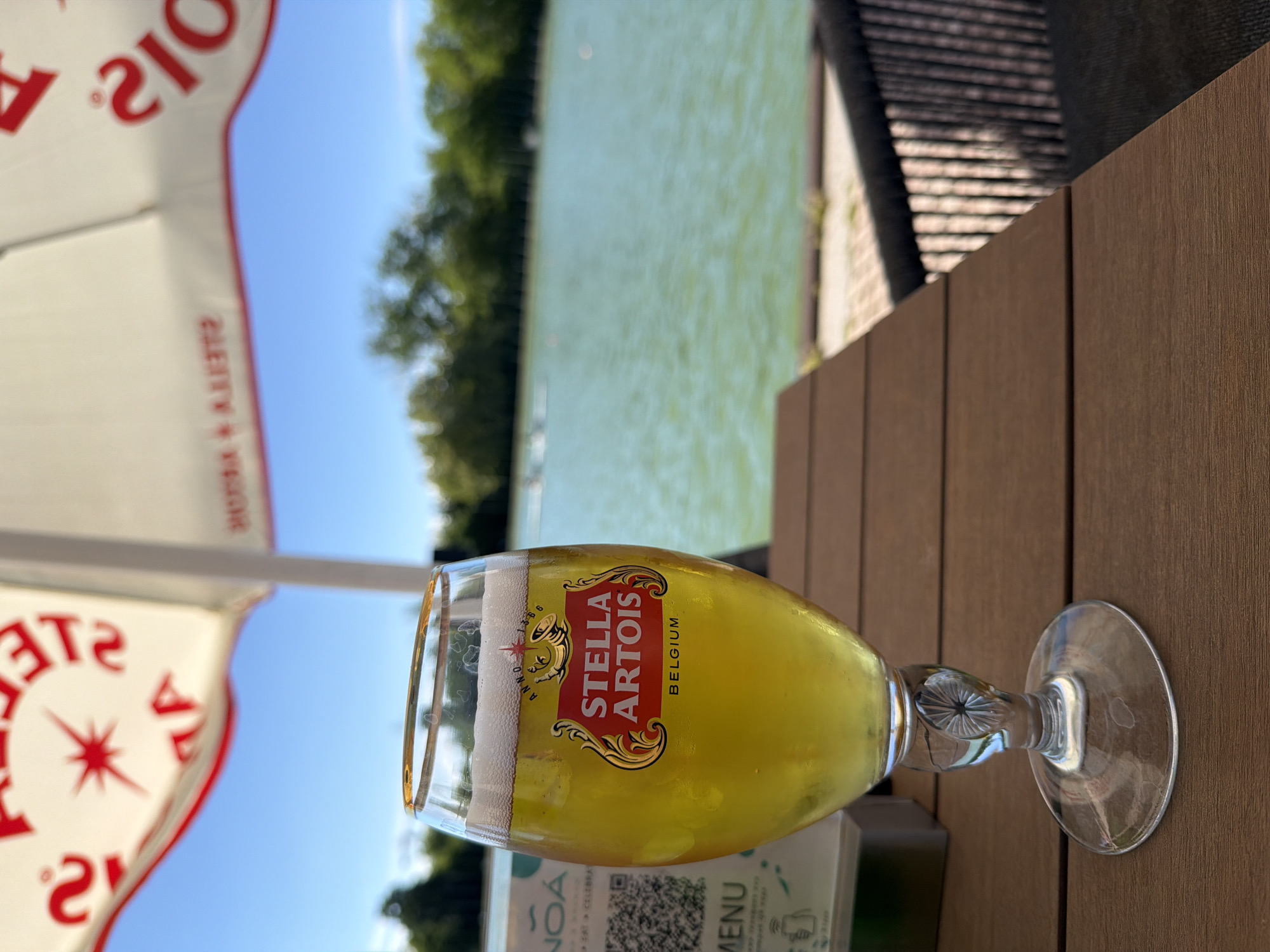 A glass of Stella Artois beer sits on a wooden table next to a menu. An umbrella with the Stella Artois logo is partially visible on the left. The background includes a view of water and trees. The photo appears to be taken at an outdoor location in Plovdiv, Bulgaria.