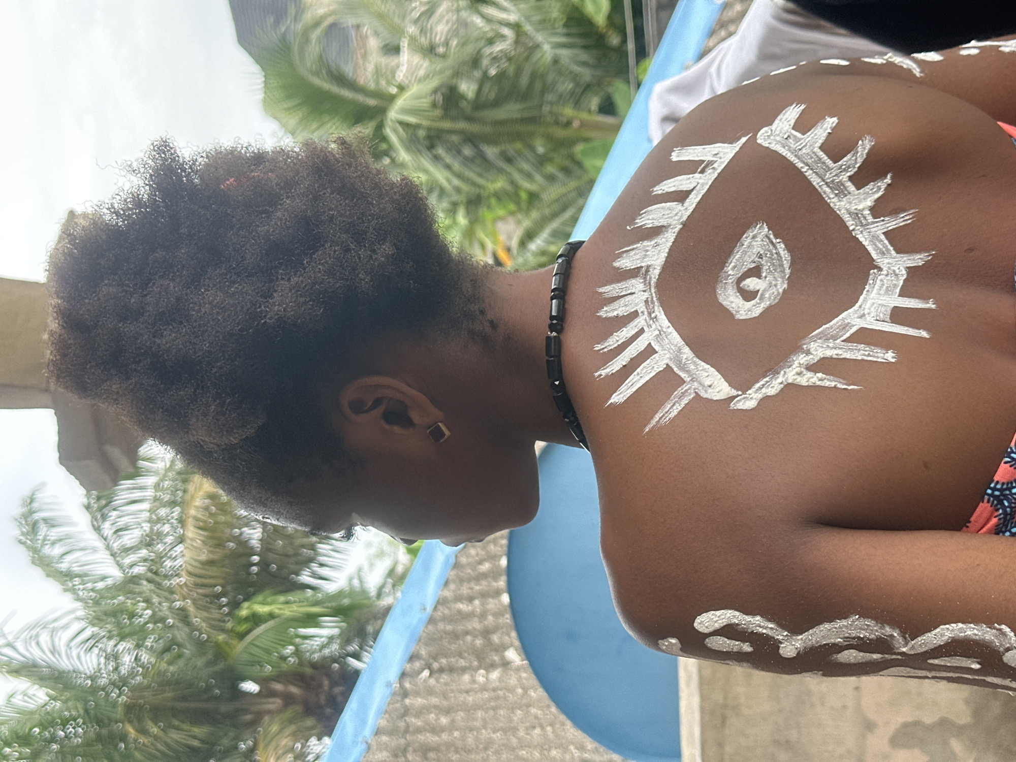 In Badagry, Nigeria, a young Black person is shown from behind, facing away from the camera, with their head turned to the left. They have dark, curly hair styled in an afro and are wearing a black beaded necklace. Their back and upper arm are adorned with white body paint. The paint on their back is a stylized eye with lashes, and swirling patterns are painted on their arm. They are wearing a patterned top. The background features lush green palm trees, a grey concrete wall, and a bright blue structure. The lighting suggests it is daytime, with the sky appearing overcast.