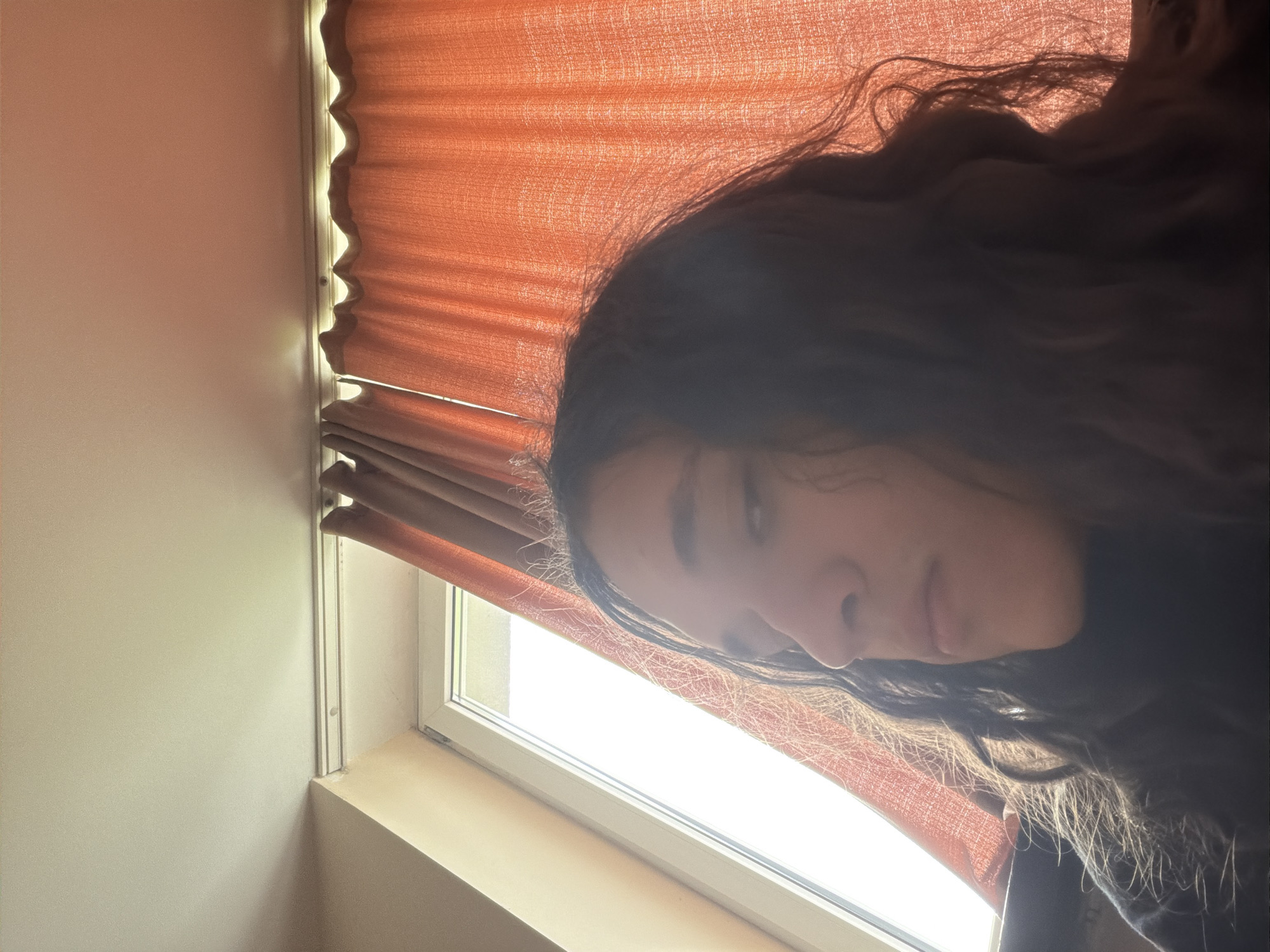 A person with long, dark, curly hair is positioned in front of a window covered by a red-orange shade. The window frame is white and there is a light-colored wall to the left. The person's face is partially visible, angled downwards towards the camera. The specific location in Plovdiv, Bulgaria is not identifiable from the image.