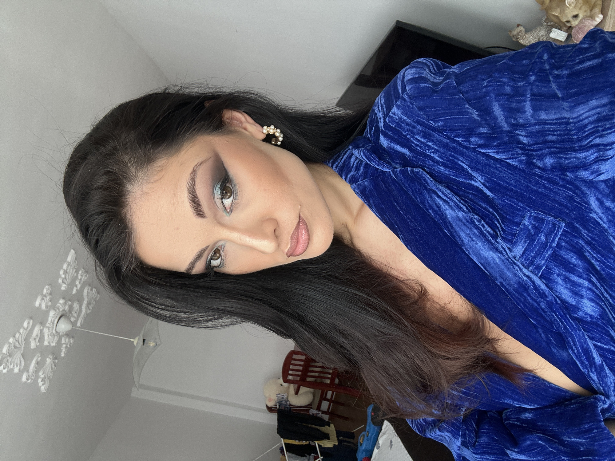 A woman is seen in a medium shot, angled slightly to the left. She has dark hair and is wearing a bright blue, textured blouse or dress. Her makeup is noticeable, with pronounced eye makeup. She is wearing pearl earrings.
The background shows parts of a room: a white wall with decorative ceiling features and a hanging light fixture, a television screen, and an indistinct assortment of household items including a small red chair.