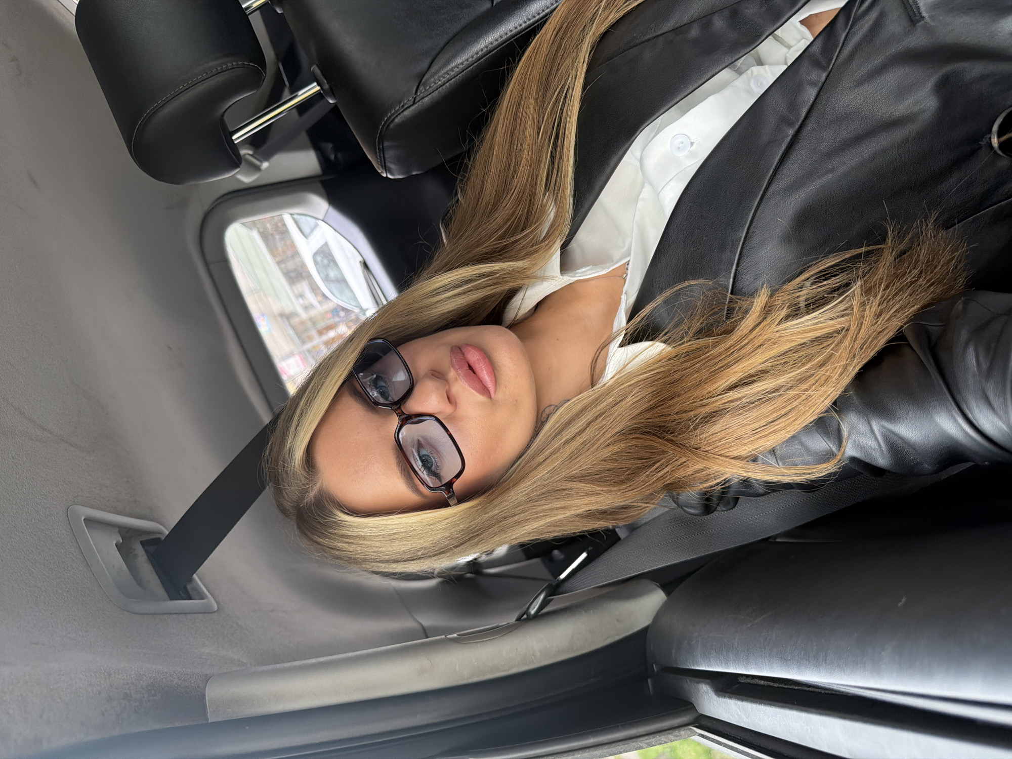The image shows a woman inside a vehicle. She has long blonde hair, wears dark-framed glasses, a white collared shirt, and a black leather jacket. The interior of the car is visible, including the leather seats, seatbelt, and the car's window. The location, based on the visual context provided, cannot be confirmed as Berlin, Germany without additional details within the image.