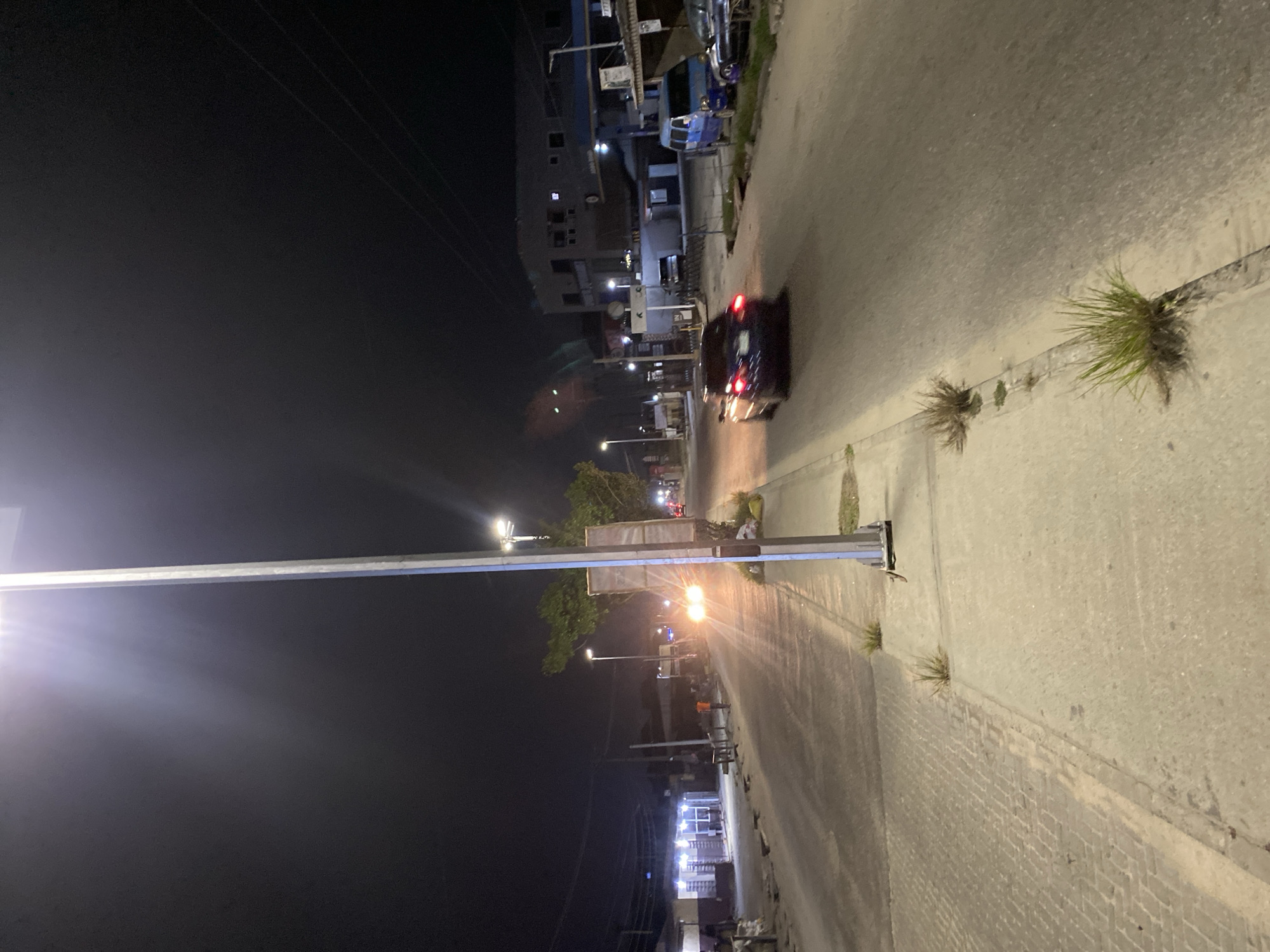 The image depicts a nighttime scene in Ikorodu, Nigeria, showing a street lit by streetlights. A car with illuminated taillights drives along the road. Sidewalks are visible on the sides of the street. Buildings and vegetation are present. There are no visible people in the image.