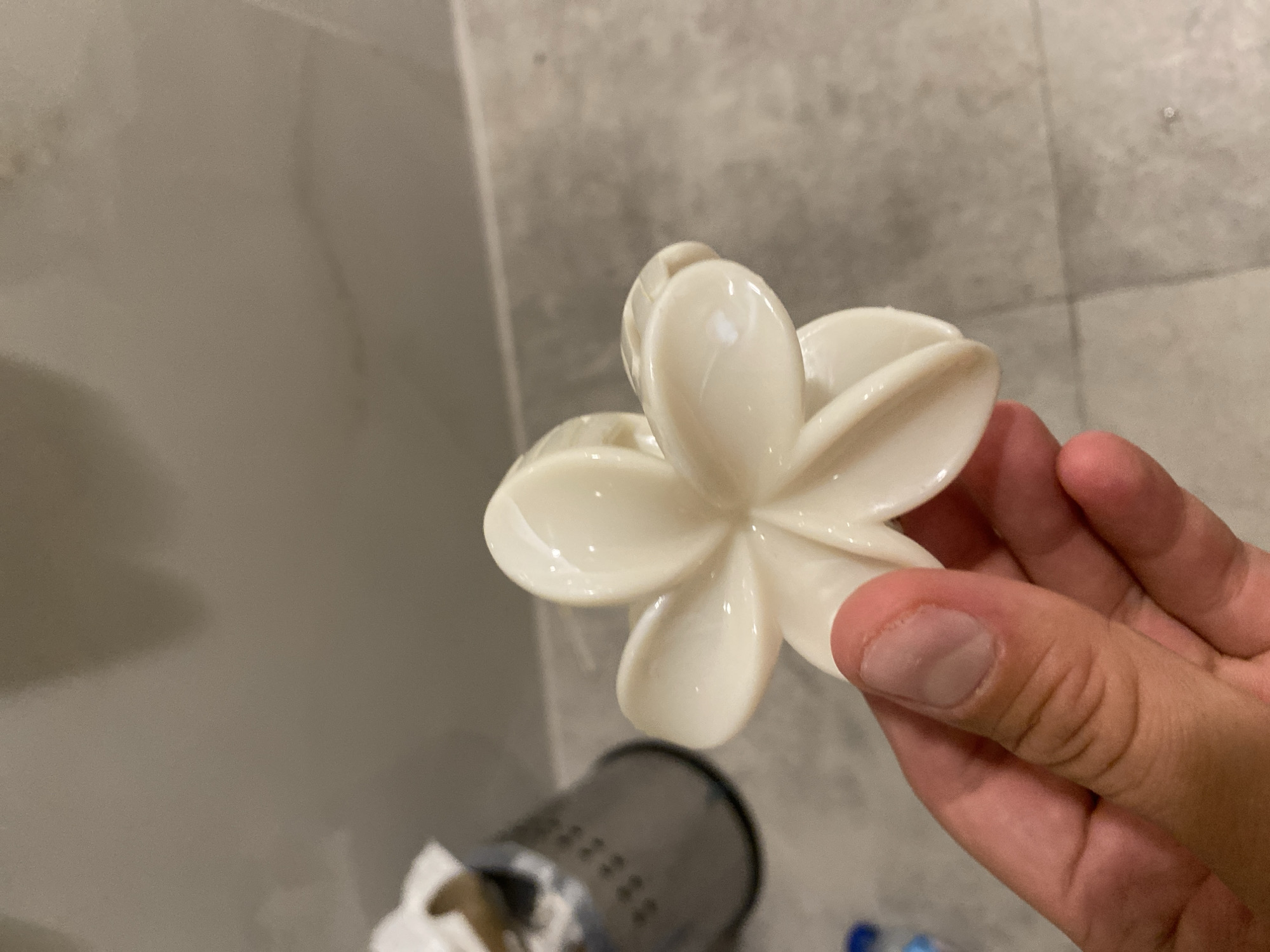 A person's hand is holding a creamy white, five-petal flower-shaped hair clip against a background of tiled flooring and a wall. In the blurred background, a silver cylindrical object with perforations is visible, possibly a trash can. The lighting is artificial and casts shadows, suggesting an indoor environment. The scene is devoid of any specific urban or natural elements that would indicate its location in Aksu, Turkey.