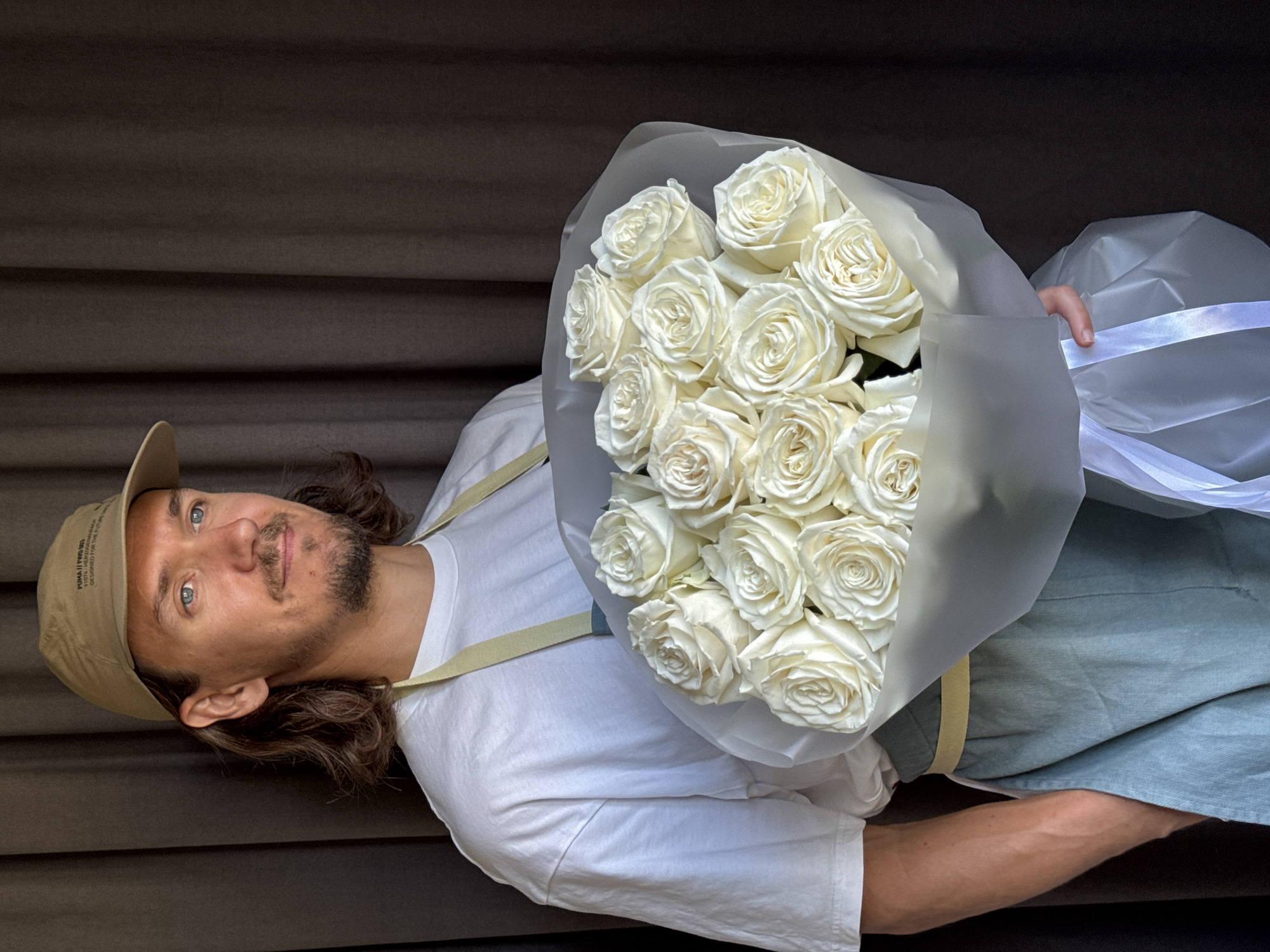 A man with long brown hair, a beard, and blue eyes, wearing a tan baseball cap, a white t-shirt, and a light green apron, stands against a dark brown curtain and holds a large bouquet of white roses wrapped in white paper and tied with white ribbon. The man is looking to his right. The bouquet is positioned in front of his chest. The scene is indoors. The location of this image is not specified in the provided information, thus it cannot be determined to be in Izhevsk, Russia.