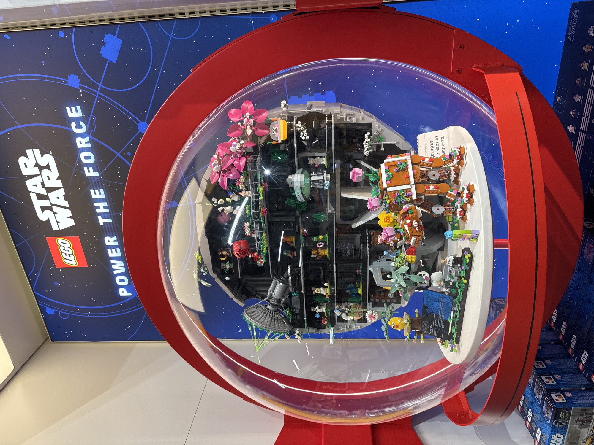 The image captures an elaborate, customized LEGO display presented within a clear, spherical enclosure framed by a prominent red ring. The central subject is a large, spherical LEGO model resembling a Death Star, but it is heavily adorned with natural and whimsical elements. Numerous pink and white LEGO flowers, miniature trees, various small LEGO animals, and even a gingerbread house-like structure are integrated into the design, creating a unique blend of sci-fi and fantasy. Several LEGO minifigures are also visible within the intricate interior sections of the model, alongside smaller spaceship-like builds.
The display is situated in what appears to be a retail environment, likely a LEGO store. The background features a large dark blue wall or poster with the "STAR WARS LEGO POWER THE FORCE" logo and a starry, geometric design. To the right, stacks of LEGO Star Wars set boxes are visible, reinforcing the theme. The overall setup is a static exhibition designed to showcase creative LEGO building.
Visible text includes "STAR WARS", "LEGO", and "POWER THE FORCE" on the background graphic. Inside the display, a small white sign is visible, reading "Death Star" with additional, largely illegible English text below it. Another partially visible sign written in Cyrillic script states "Декорът е част от комплекта," which translates to "The decor is part of the set." The scene is brightly lit, consistent with an indoor retail setting. No people are present in the image.