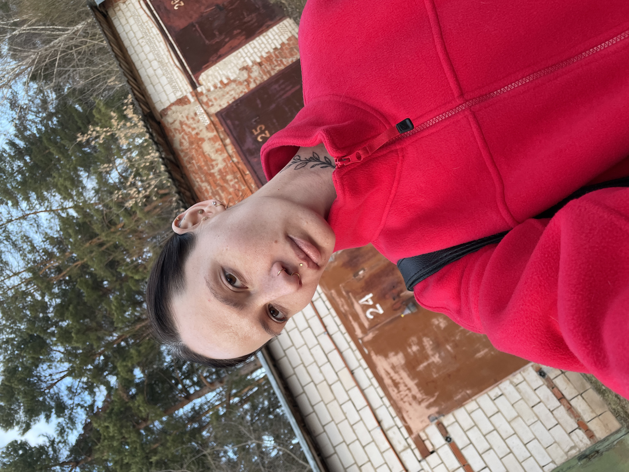 A person with short dark hair, multiple ear piercings, a nose ring, and a leaf tattoo on their neck is wearing a red fleece jacket. The background shows a brick building with numbered garage doors (24, 25, and 26 are visible). Trees and sky are seen in the background. The photo appears to be taken outdoors in Oktyabr'skii, Russia.