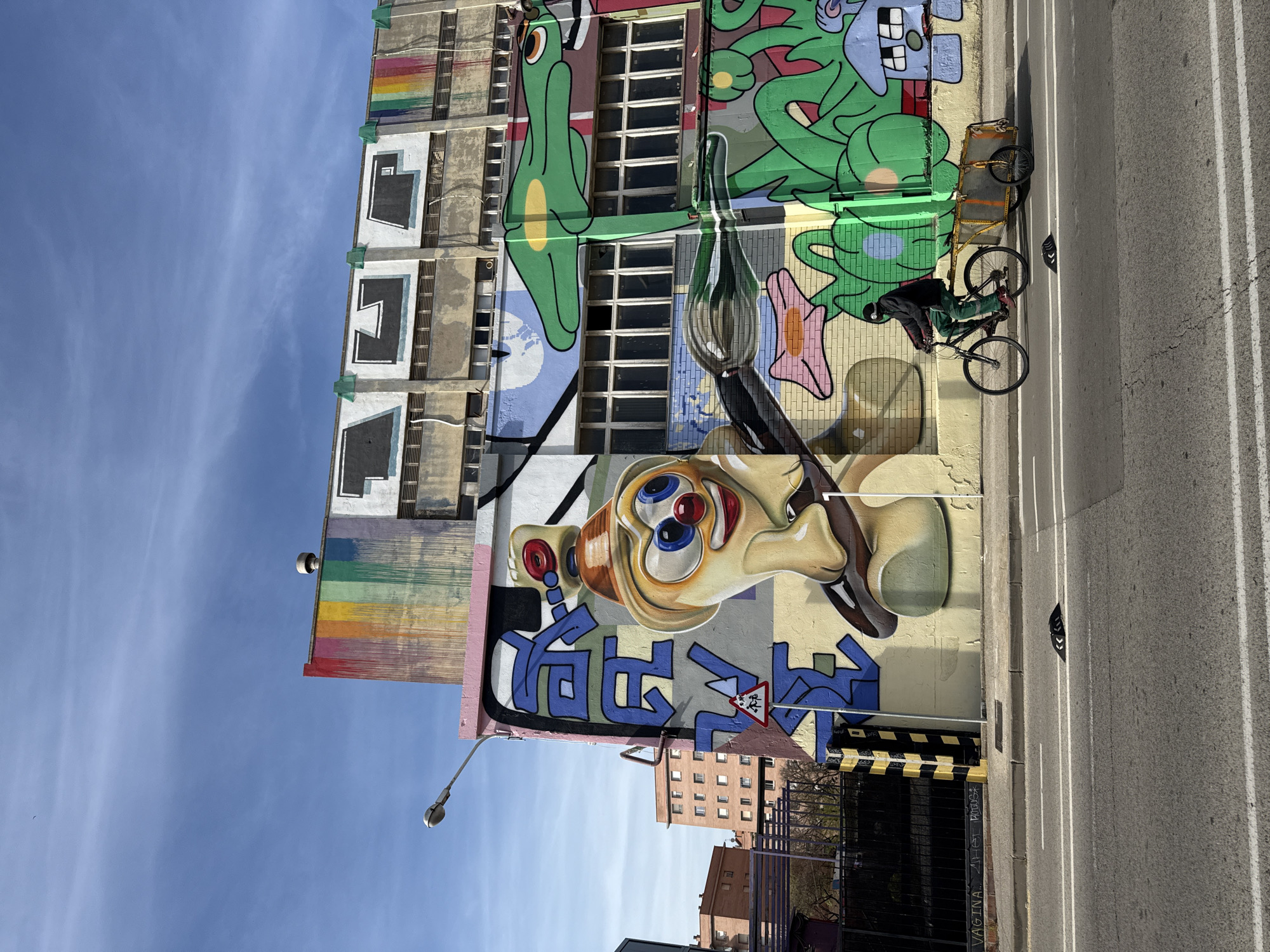 An eye-level shot depicts a building in Barcelona, Spain, covered with colorful graffiti art. Prominently featured is a large cartoon face with bulging eyes and a bulbous nose. Above and around the face are various abstract shapes and figures in greens, blues, and yellows. A person on a bicycle with a trailer is positioned to the right of the mural along a city street. The street is asphalt and has white lane dividers. A street lamp stands near the building. Other buildings are visible in the background. The sky is clear and blue.