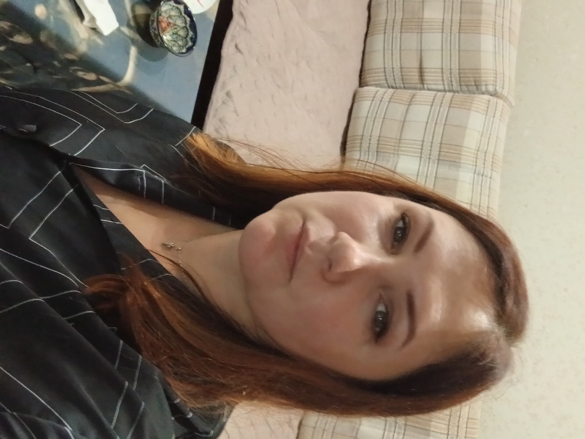 A medium shot photo shows a woman with reddish-brown hair and light skin. She is wearing a black collared shirt with a white geometric pattern. Her hair is shoulder-length and parted on the side. She has on light makeup, including eyeliner and mascara. A thin silver necklace with a small pendant is visible around her neck. The background shows a sofa with beige plaid cushions and a light-colored blanket. To the left of the woman, on a dark-colored table, is a small blue and white patterned bowl and a white napkin. The overall lighting appears to be artificial.