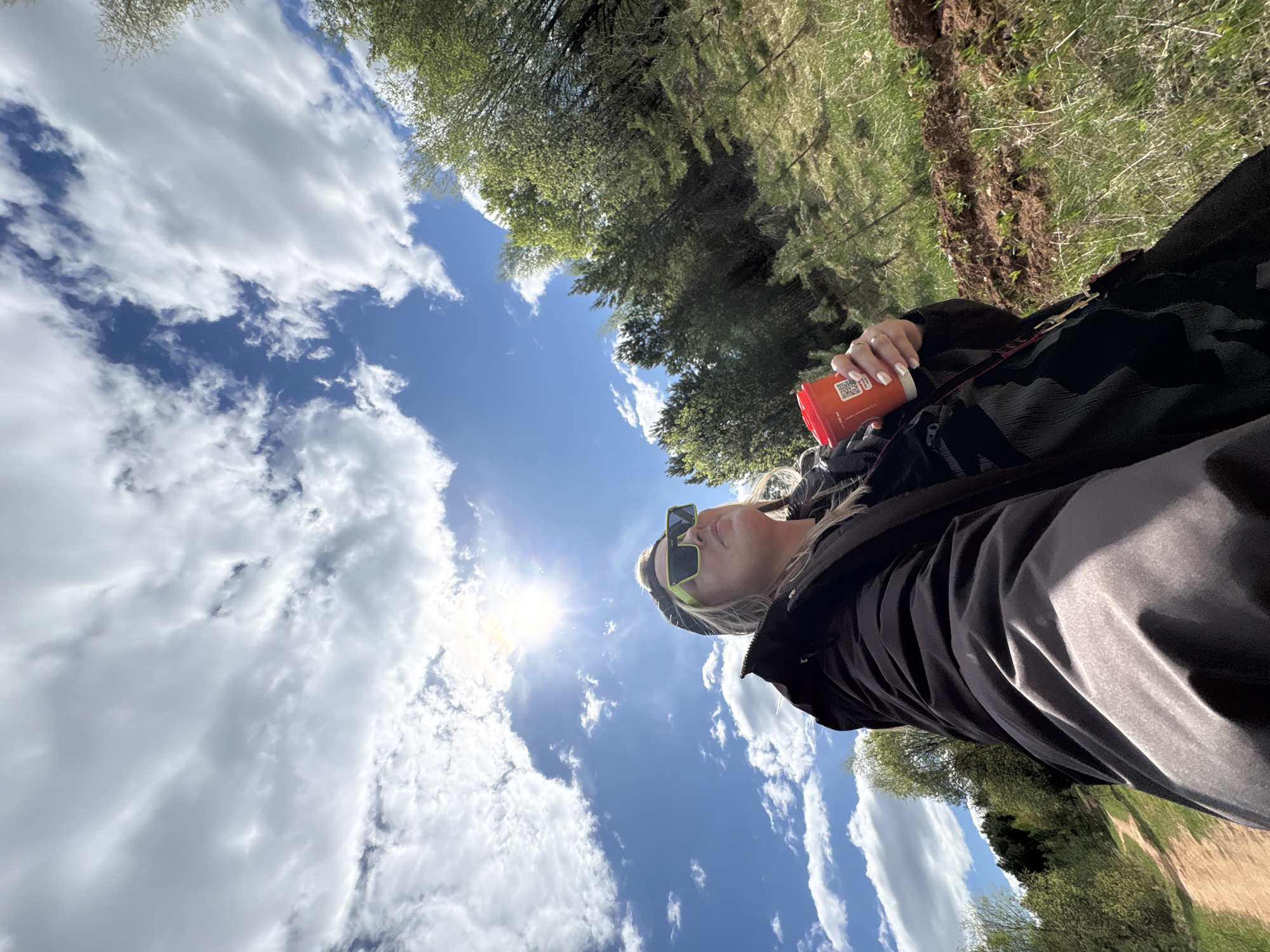 The photograph is taken from a low angle, looking up at a person lying on the ground. The person is wearing dark clothing and neon green sunglasses, and holding a red beverage container in their hand. The background includes a bright blue sky with fluffy white clouds and trees. Based on the environment and foliage, it is likely taken near a wooded area or park in the city of Марковский, Russia.