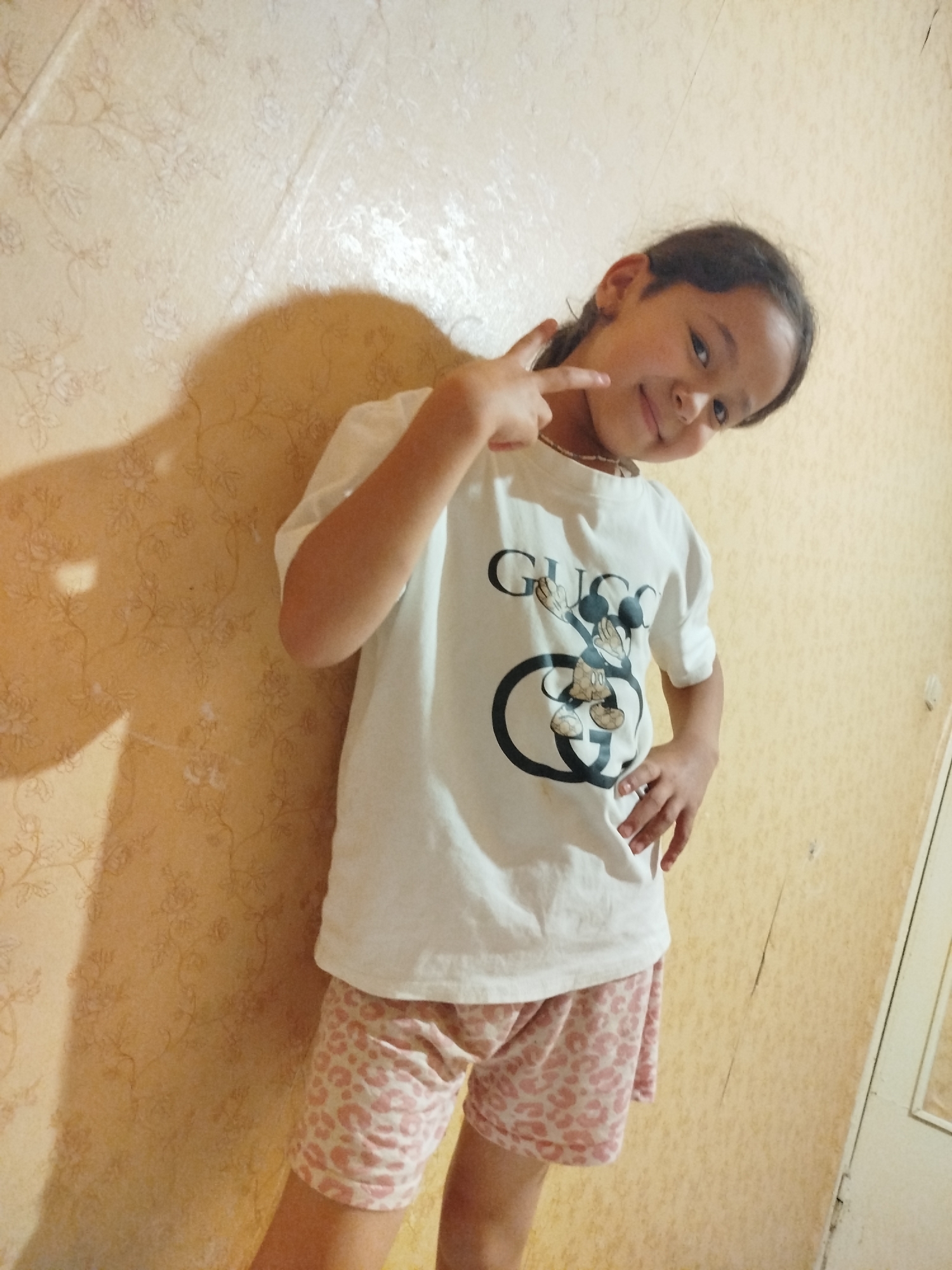 A young girl, likely of East Asian descent, stands against a textured, yellow wall with a faint floral pattern. She is wearing a white t-shirt with a graphic of Mickey Mouse within a Gucci logo, and light pink shorts with a leopard print. Her hair is dark brown and styled in a braid. She is looking towards the camera with a slight smile and her head tilted. Her right hand is raised, forming a peace sign, while her left hand rests on her hip. A shadow of her right arm is cast on the wall behind her. The lighting suggests an indoor setting, possibly during the daytime. The girl appears cheerful and playful. No specific time of day or weather conditions can be determined. There is text on her shirt, reading "GUCCI" and featuring the Mickey Mouse graphic. The location is identified as Ivanovo, Russia.