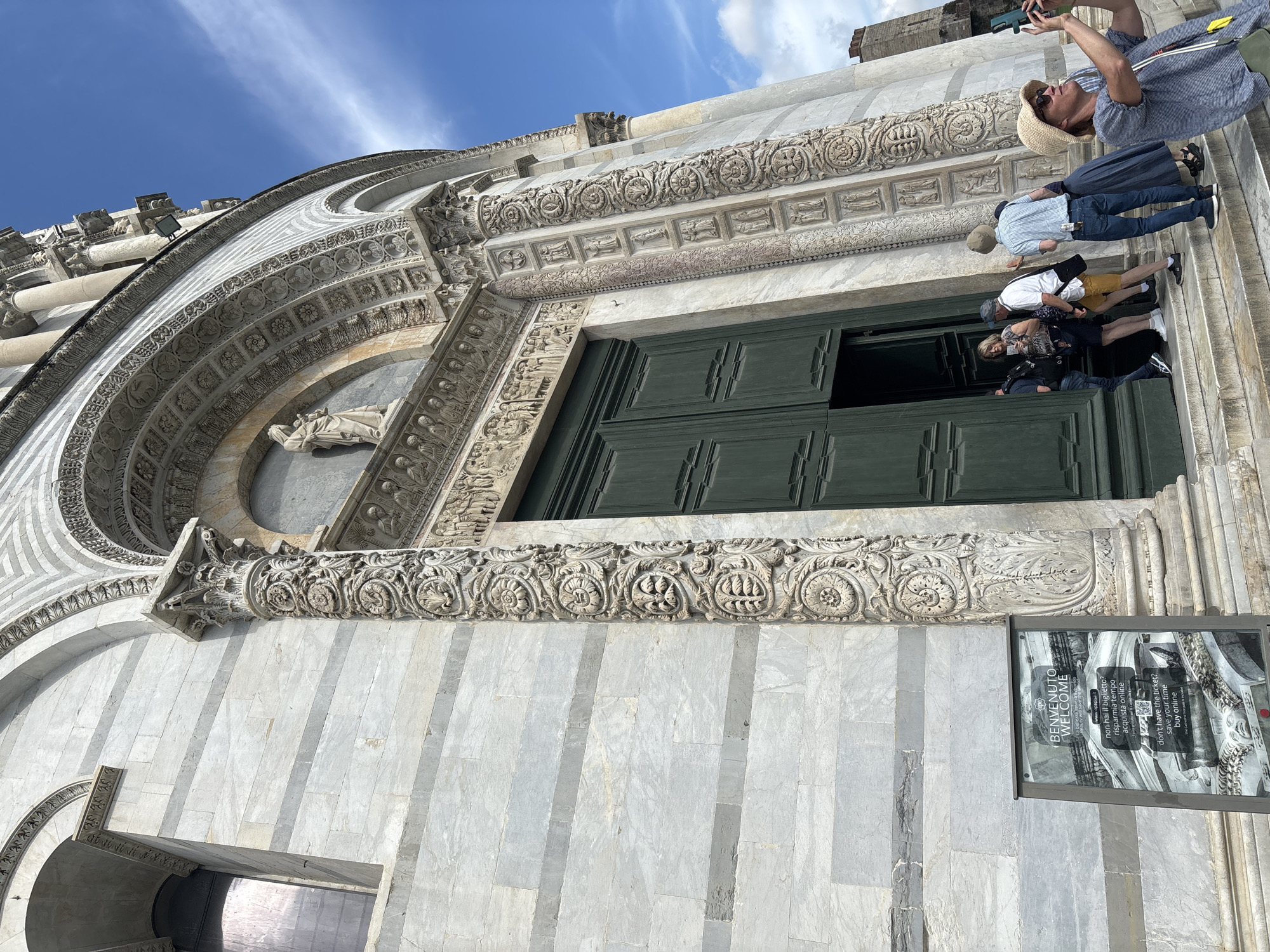 The image shows a section of the Pisa Cathedral, located in Pisa, Italy. The view includes a large ornate entranceway with closed green doors. Above the doors are carved decorative details and a statue of a figure. A group of tourists are standing on the steps leading to the entrance, with one taking a photo. To the side of the entrance, there is a sign with "Benvenuto, Welcome" printed on it, encouraging visitors to buy tickets online. The architecture is mainly white marble with grey stripes. The sky in the background is blue with some clouds.
