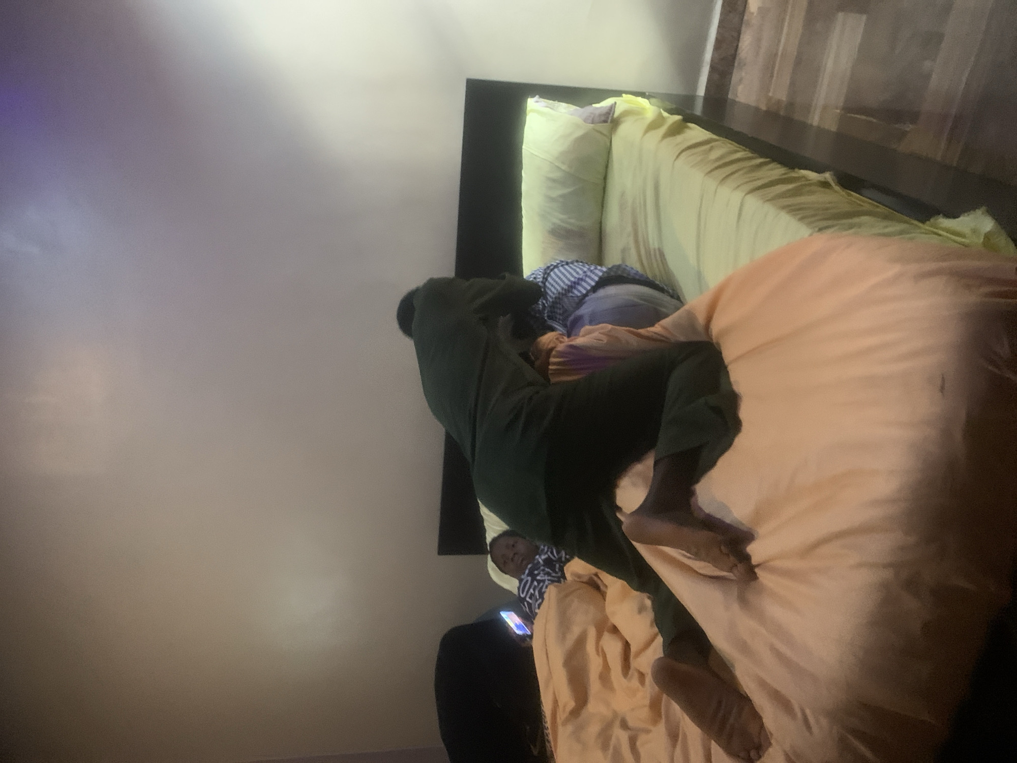 In a bedroom in Nkpor, Nigeria, a person is on all fours on a bed, facing away from the camera. Their dark clothing contrasts with the pale orange duvet and pale yellow fitted sheet. To their left, a person lies on their back, propped up on a pillow, looking at a phone with a bright screen. The room has beige walls and wooden floors. A dark headboard is visible behind the bed.