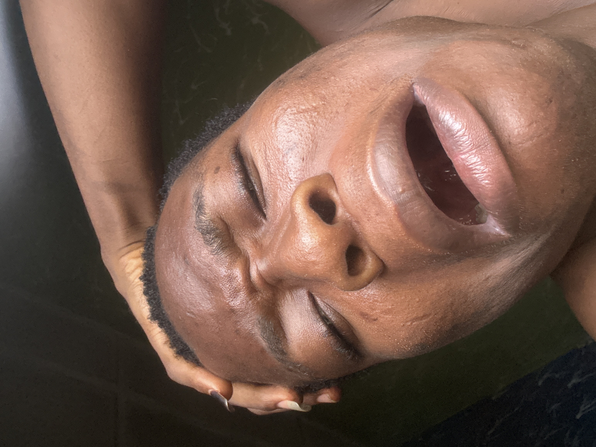 A close-up, tilted perspective of a dark-skinned person with closed eyes and an open mouth is shown. The person's face is illuminated, revealing pores and slight wrinkles. The hands of the person are seen supporting the head, with the fingernails of the supporting hand being long and polished. The background is dark and out of focus. The location is unknown.