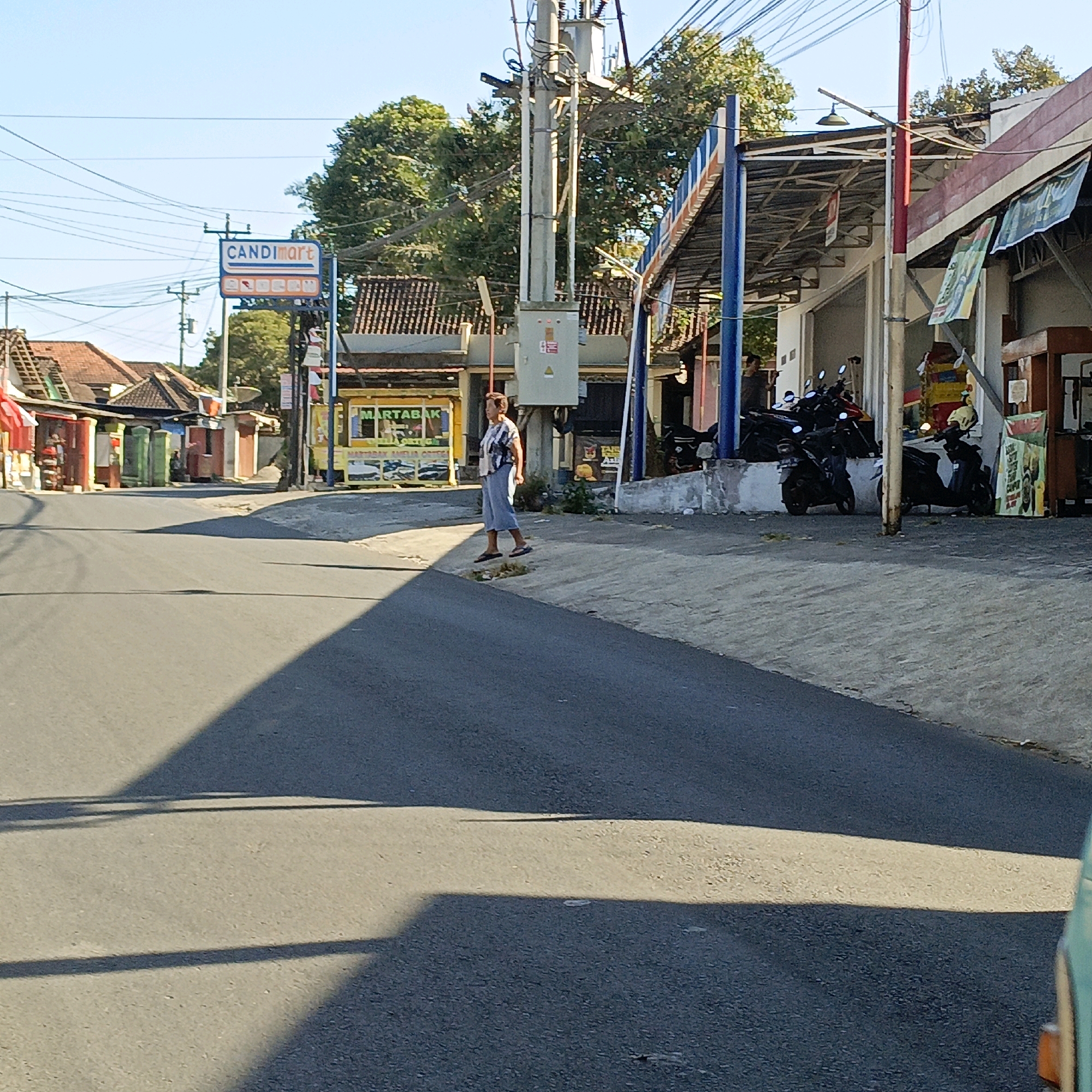 A street scene in Candirejo, Indonesia, features a lone pedestrian walking on a sidewalk next to a road. The pedestrian, a woman wearing a patterned shirt and light-colored pants, is positioned mid-stride, with her left foot off the ground. She is walking towards the right side of the frame, in front of a row of storefronts. To her left, a "CANDImart" sign is visible, and several other shops and buildings line the street. Motorcycles are parked in front of some of the establishments. Overhead, power lines crisscross the sky, and trees are visible in the background. The road is paved and appears to be a typical urban street, with shadows cast by the buildings and power poles. The overall impression is of a quiet, sunny day in a commercial area.