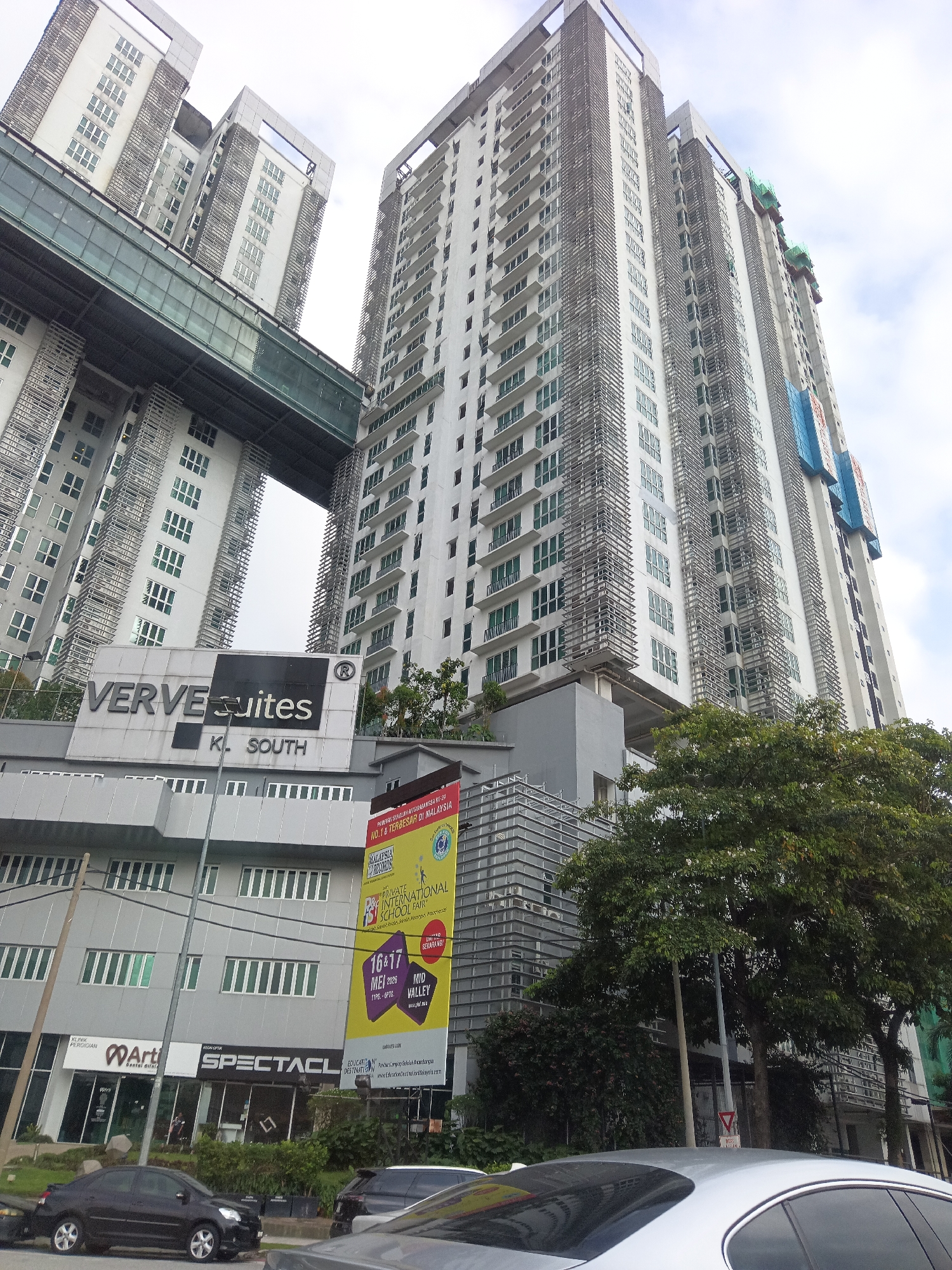 This is a low-angle, eye-level, full shot of the Verve Suites KL South residential complex in Kuala Lumpur, Malaysia. The complex features two prominent, tall, white apartment buildings with a connecting skybridge. The building on the left is partially obscured by the building on the right. The skybridge, constructed with glass and metal, connects the two buildings at a high elevation. The apartment buildings have numerous windows and external decorative elements. In front of the buildings, there is a lower-rise structure with various businesses, including "Artis Dental Clinic" and "SPECTACL." A large yellow banner advertising a "Private International School Fair" is displayed on the front of the lower building. Several trees and lush greenery surround the base of the complex. The sky is overcast. Cars are parked or driving in the foreground, including a black sedan and a silver sedan. The image is taken on a bright day, with the sun diffused by clouds.