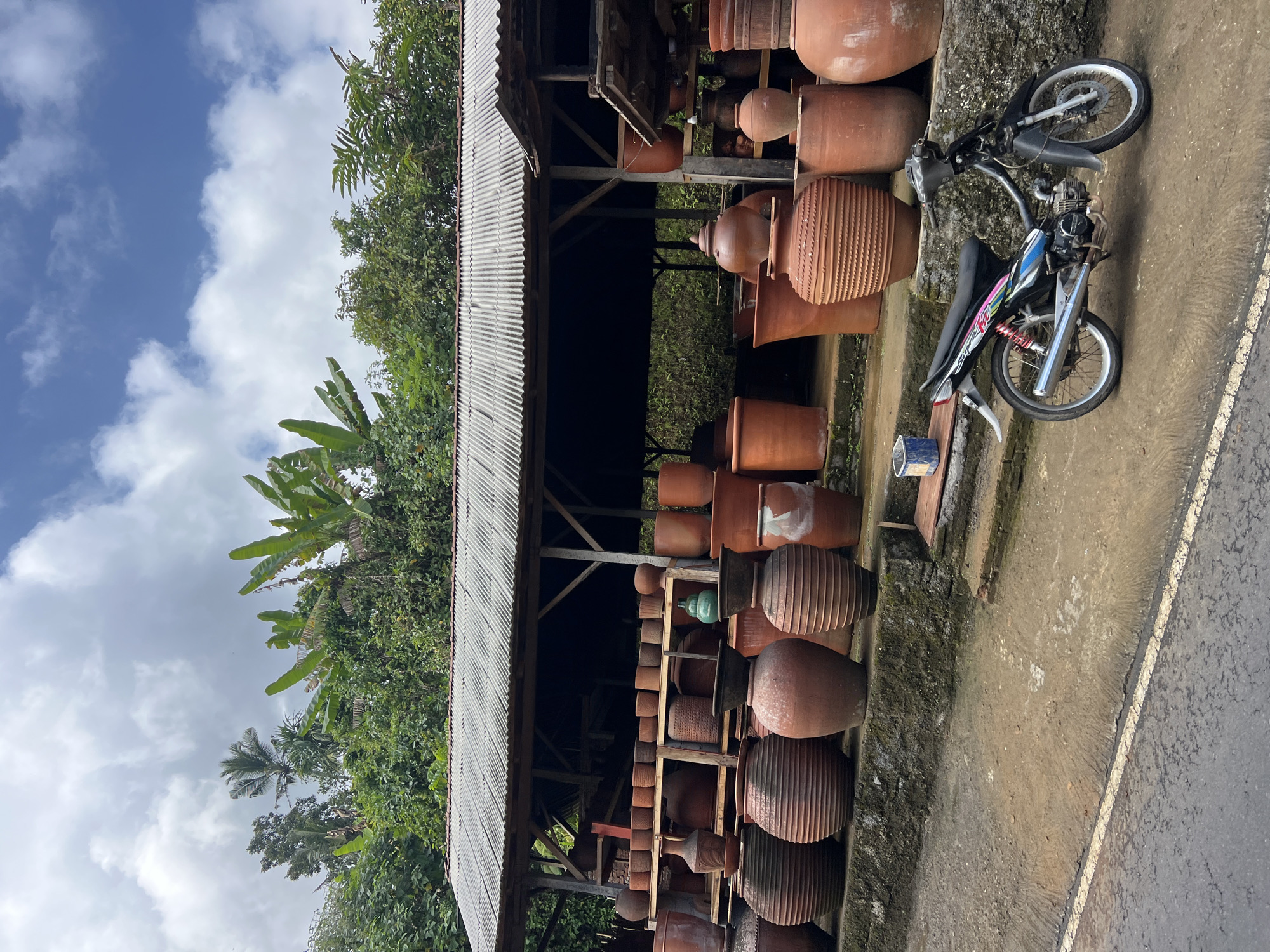 The image depicts an outdoor pottery stall in Tembuku, Indonesia, showcasing a wide array of terracotta pots of various sizes and designs. A corrugated tin roof supported by wooden posts provides shelter for the display. The pots are arranged on shelves and a low stone wall, with some larger pieces placed on the ground in front. Lush green foliage and trees form a backdrop, suggesting a rural or semi-rural setting. A black motorcycle is parked in front of the stall, facing right, with its kickstand down. The ground is a mix of dirt and paved areas, with a white line marking a road edge in the foreground. The sky is cloudy with patches of blue.