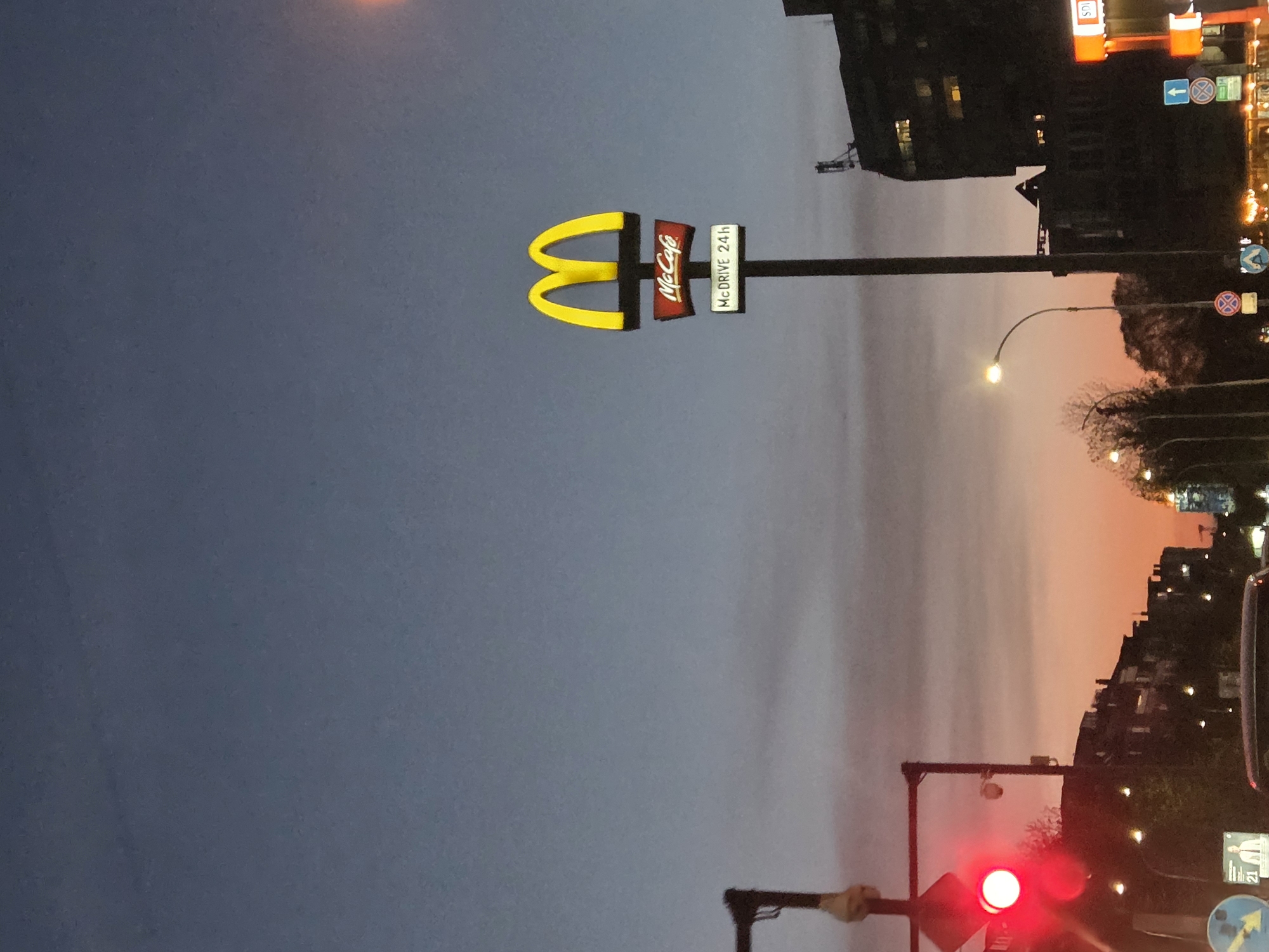Captured at dusk, the image features a McDonald's sign in Varna, Bulgaria. The sign, displaying the golden arches, McCafe logo, and "McDrive 24h" inscription, stands prominently against a twilight sky tinged with orange hues on the horizon. Below the sign, a traffic light glows red. Buildings line the background, marked by the lights of the city at night. The image is taken from a slightly low angle, possibly from a vehicle at a standstill.