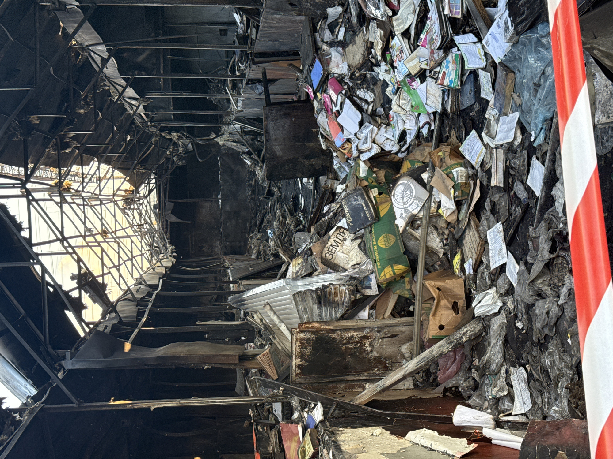 The image shows the interior of a damaged building, possibly a market or warehouse, in Kyiv, Ukraine, following a fire. The roof structure is severely damaged, with exposed beams and torn insulation. Debris, including burnt paper, books, and corrugated metal, fills the central area of the building. Several cardboard boxes are visible, some collapsed and others intact, containing items that appear to be books or documents. In the foreground, a red and white caution tape is stretched across the bottom of the frame, indicating a restricted area. The environment is dark and appears to be filled with smoke residue and ash.