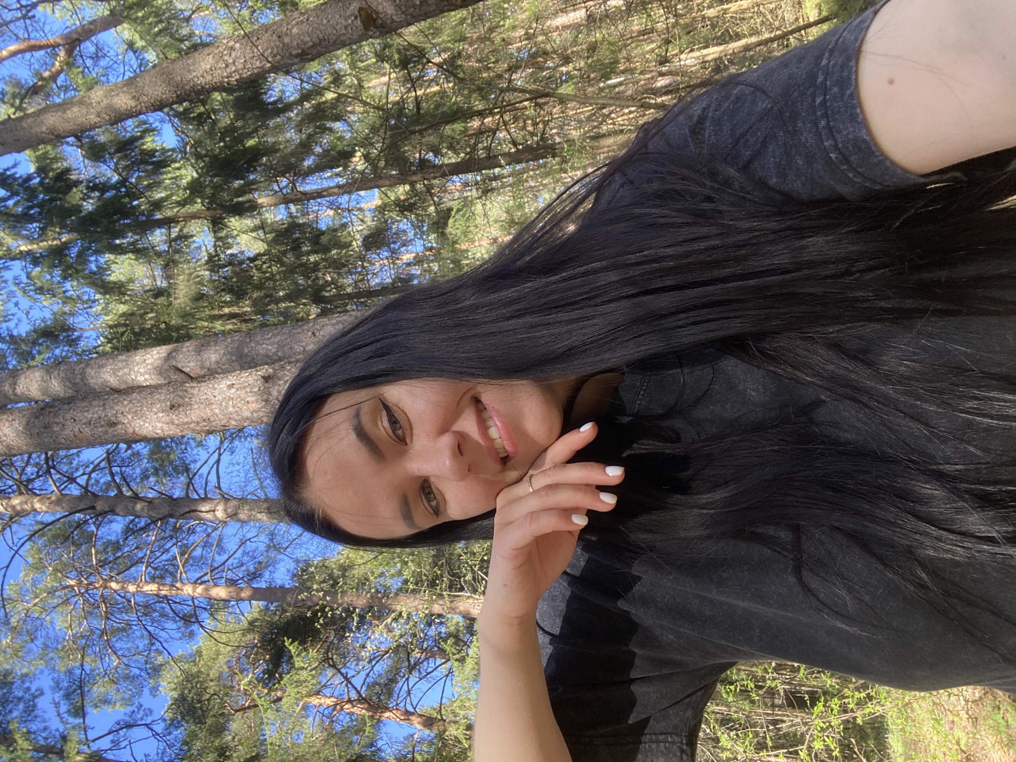 A woman with long black hair, wearing a dark gray shirt, is posing for a selfie. She is smiling and resting her face on her hand, which has white nail polish and a gold ring. The background shows a forest with tall trees and a blue sky visible through the branches. Based on the context, the location is likely a forest or park within the city of Perm, Russia.