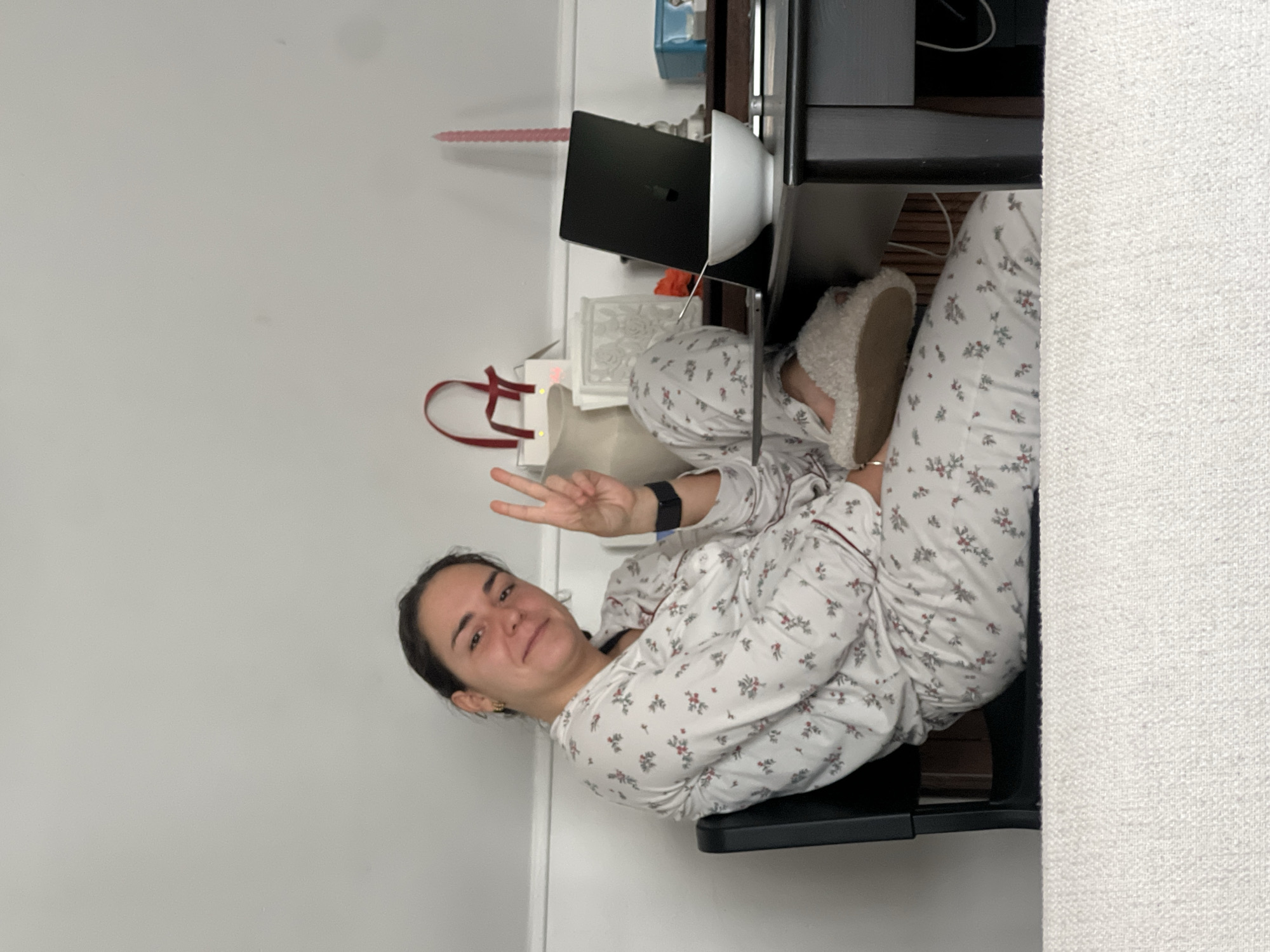 A woman in pajamas is sitting at a desk in a room with a white wall and a light-colored couch. She is giving a peace sign and smiling. There is a laptop on the desk in front of her, as well as other items such as a red bag and a candle. The location is not identifiable as Paris, France.