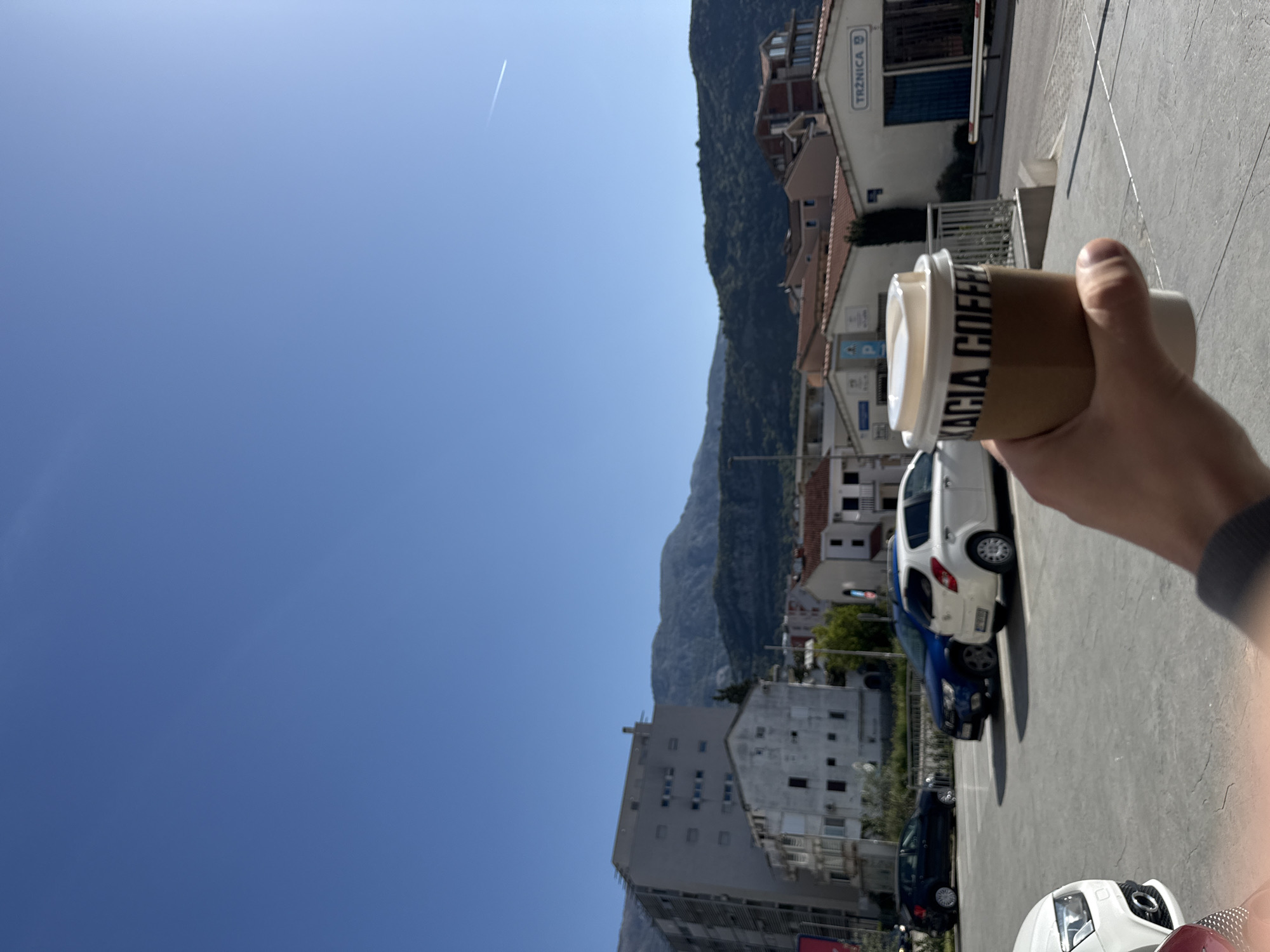 The image is taken outdoors on a sunny day, showing a person holding a cup of coffee labeled "Acacia Coffee." The background reveals a cityscape in Budva, Montenegro. The sky is clear blue, with a contrail from a plane visible. Mountains are present in the background, alongside buildings and trees. In the foreground, cars are parked on what appears to be a concrete parking lot. The image is rotated 90 degrees counterclockwise.