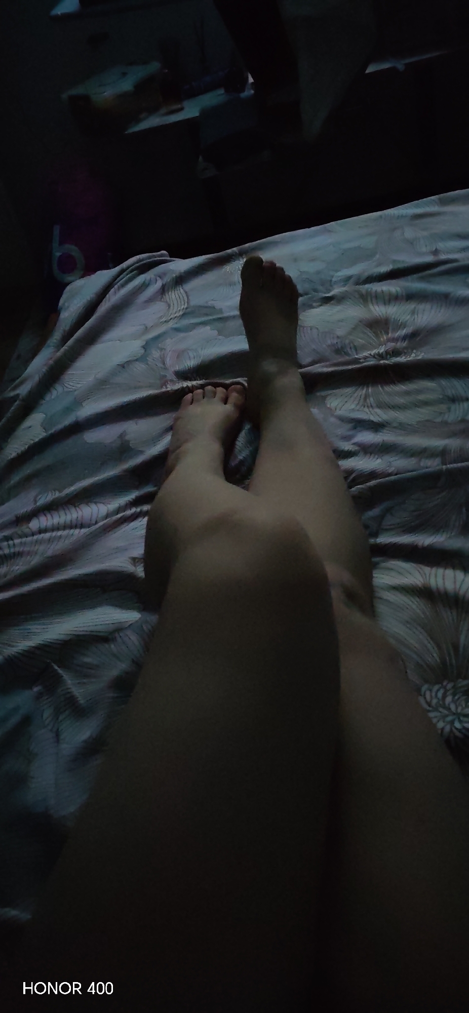 The image captures a first-person perspective of a person lying down on a bed in a dimly lit room in Noviy, Russia. The primary subjects are the person's legs and feet, resting on a patterned bedsheet. The legs are angled, with one foot partially overlapping the lower shin of the other leg. The bedsheet features a light-colored pattern, possibly floral or abstract, against a darker background, but its colors are muted due to the low light.
The background is very dark and mostly indistinct, but hints of furniture or objects like a table or dresser are visible, along with faint reflections of light sources. The overall ambiance suggests it is nighttime or a very dark indoor environment, possibly a bedroom. The activity taking place is likely resting or sleeping. A notable detail is the "HONOR 400" watermark visible in the bottom left corner, indicating the device used to capture the photo.