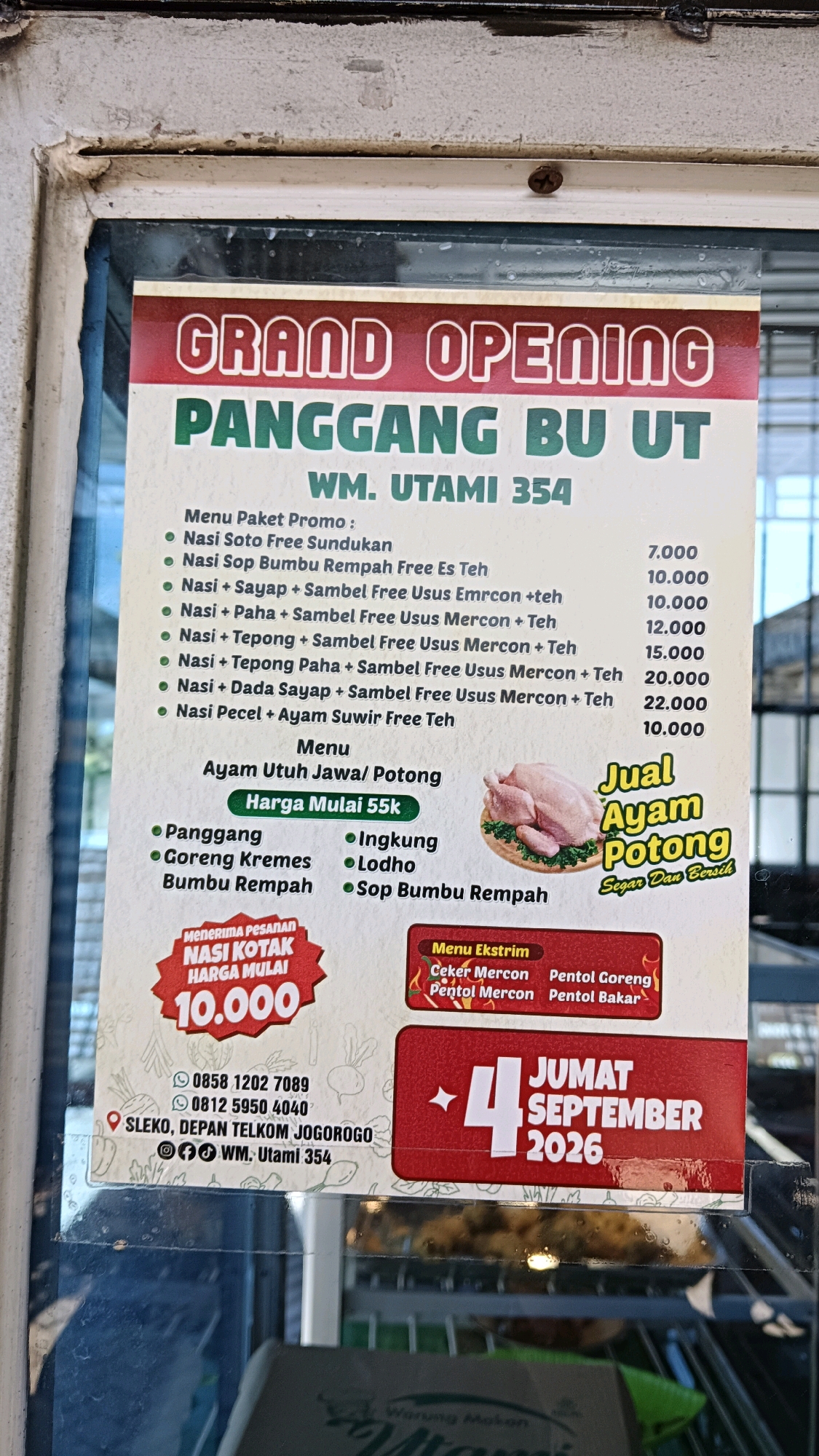 The image shows a close-up of a "Grand Opening" advertisement for "PANGGANG BU UT" (WM. UTAMI 354) located in Sleko, Depan Telkom, Jogorogo, Indonesia. The advertisement features a menu of promotional packages and individual menu items, primarily consisting of grilled chicken and rice dishes. It also highlights "Nasi Kotak Harga Mulai 10.000" (Lunch Box Price Starts at 10,000) and "Jual Ayam Potong Segar Dan Bersih" (Selling Fresh and Clean Cut Chicken). A prominent section displays "JUMAT SEPTEMBER 2026," indicating the opening date. The contact information provided includes two phone numbers: 0858 1202 7089 and 0812 5950 4040. The advertisement is displayed on a window, with reflections visible.