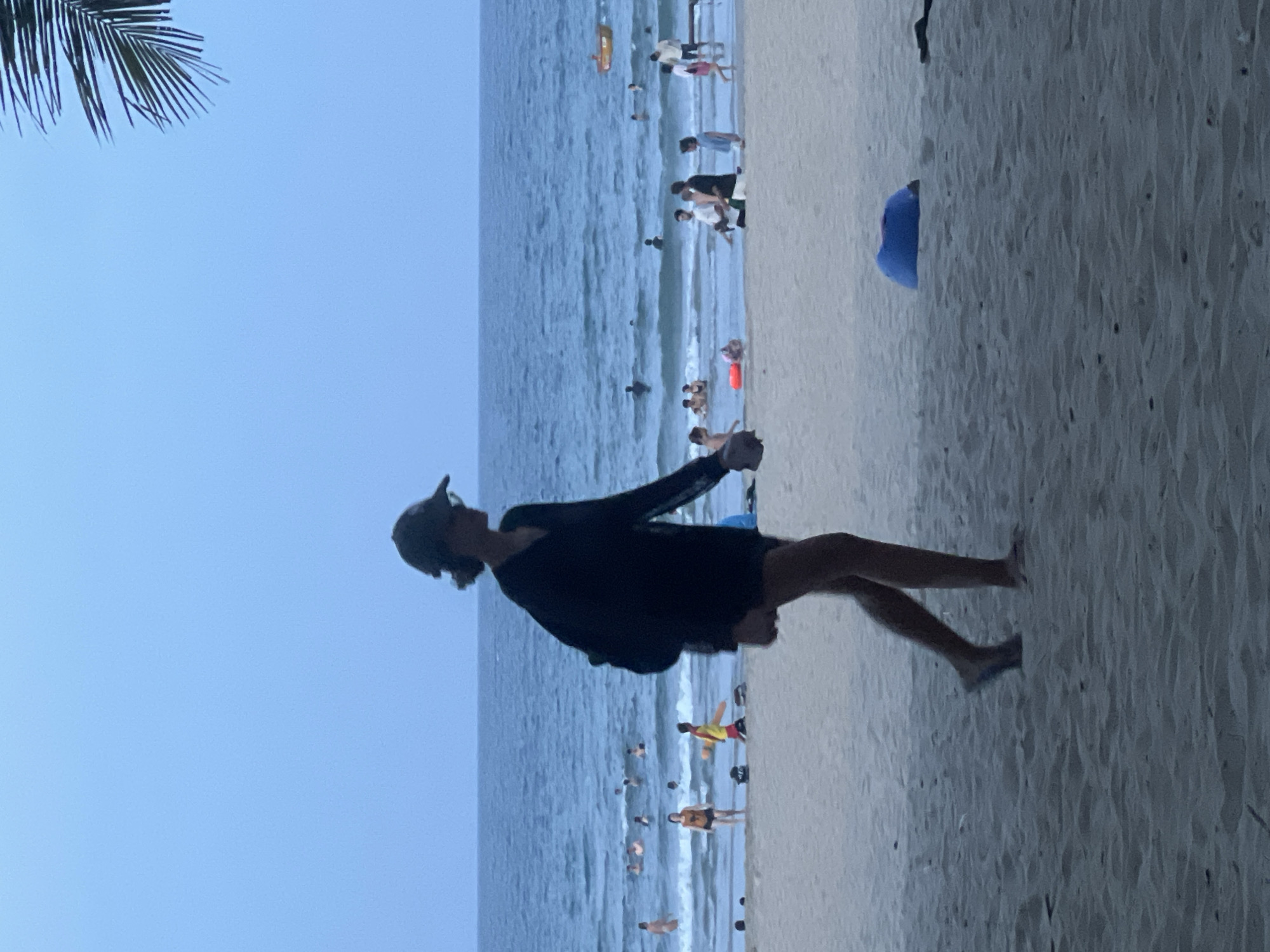 The image depicts a beach scene, likely in Da Nang, Vietnam, at dusk. A woman in silhouette, wearing a hat and a dark long-sleeved shirt with shorts, walks from left to right across the sandy foreground. Behind her, the ocean is visible with several people swimming or wading in the shallow water. Further out, a few individuals are present in the sea. The sky is a hazy blue, and a palm frond is visible in the upper right corner, suggesting a tropical location. A blue object, possibly a deflated inflatable, rests on the sand to the right of the woman. The overall atmosphere is calm and relaxed, indicative of a typical evening at a beach.