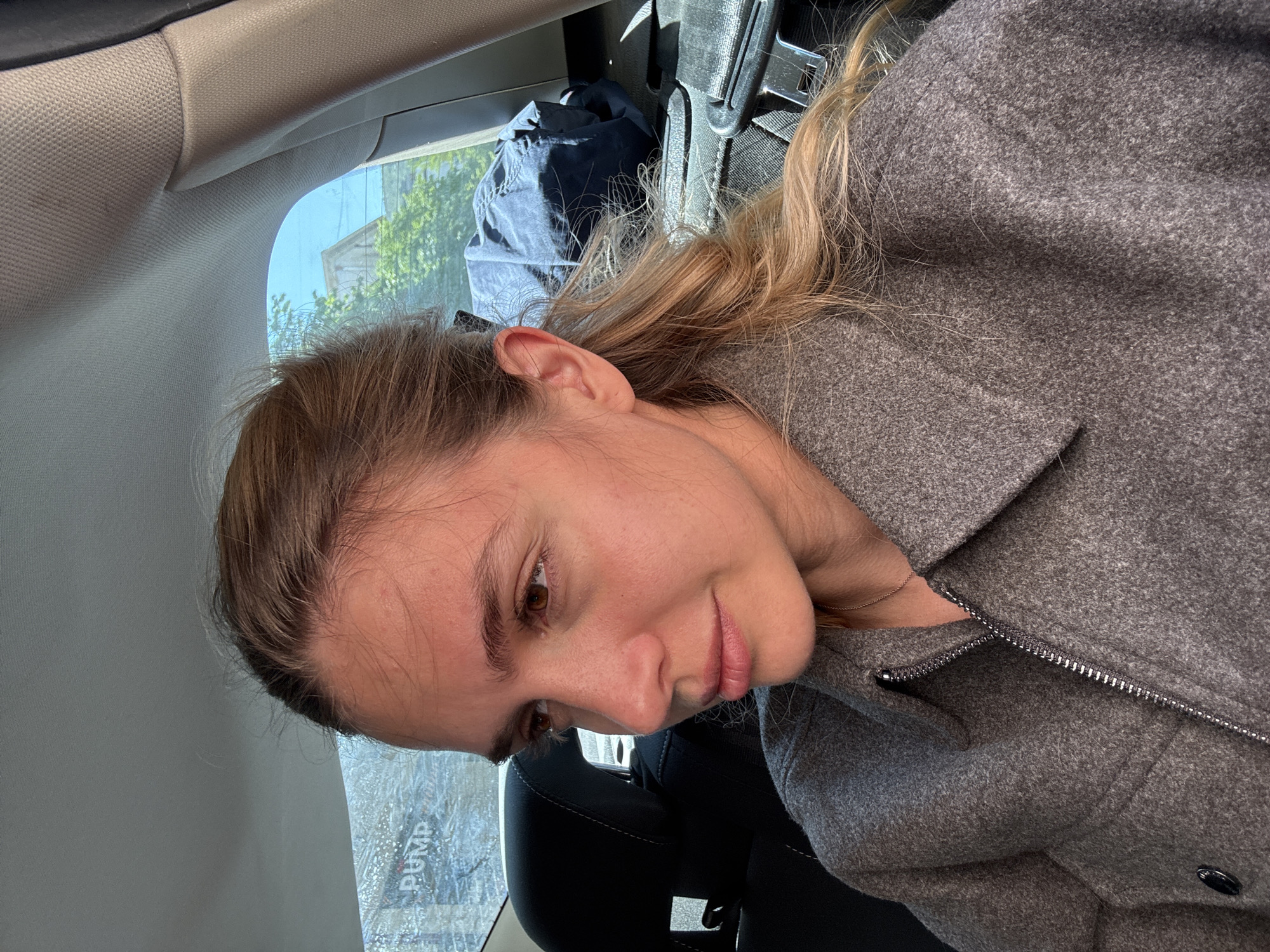 A woman with light brown hair is sitting in the back seat of a car. She is wearing a gray wool coat with a zipper. The car is parked near a building with a sign that reads "PUMP." The location appears to be in an urban area.