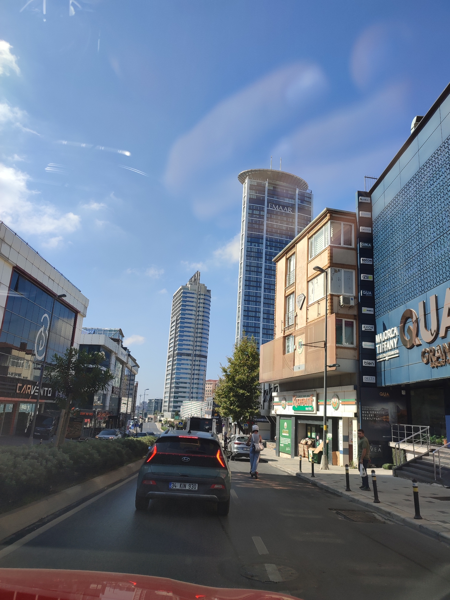 The image depicts a street scene in Üsküdar, Turkey, under a partly cloudy blue sky. The perspective is from inside a vehicle, looking forward. On the right side of the street, a modern, multi-story building with blue facade cladding and patterned geometric designs is visible. This building features a large sign with "QUA" and "MAJROCA TIFFANY" prominently displayed. Adjacent to it, a narrower, older tan-colored building with multiple windows rises up, housing various businesses indicated by vertical signs like "OXKA", "BOCCHI", and "DELTA". Further down the street, a very tall, modern skyscraper with "EMAAR" branding dominates the skyline. On the left side of the street, another modern building with a glass facade displays the "CARVENTO" logo. The street itself is paved and appears to be a main thoroughfare, with a median strip lined with bushes. Several cars are on the road, including a dark green SUV with the license plate "34 KIN 930" in the foreground. Pedestrians are visible on the sidewalk, including a person on a scooter and another individual holding a white bag. The overall impression is one of urban development with a mix of modern and older architectural styles.