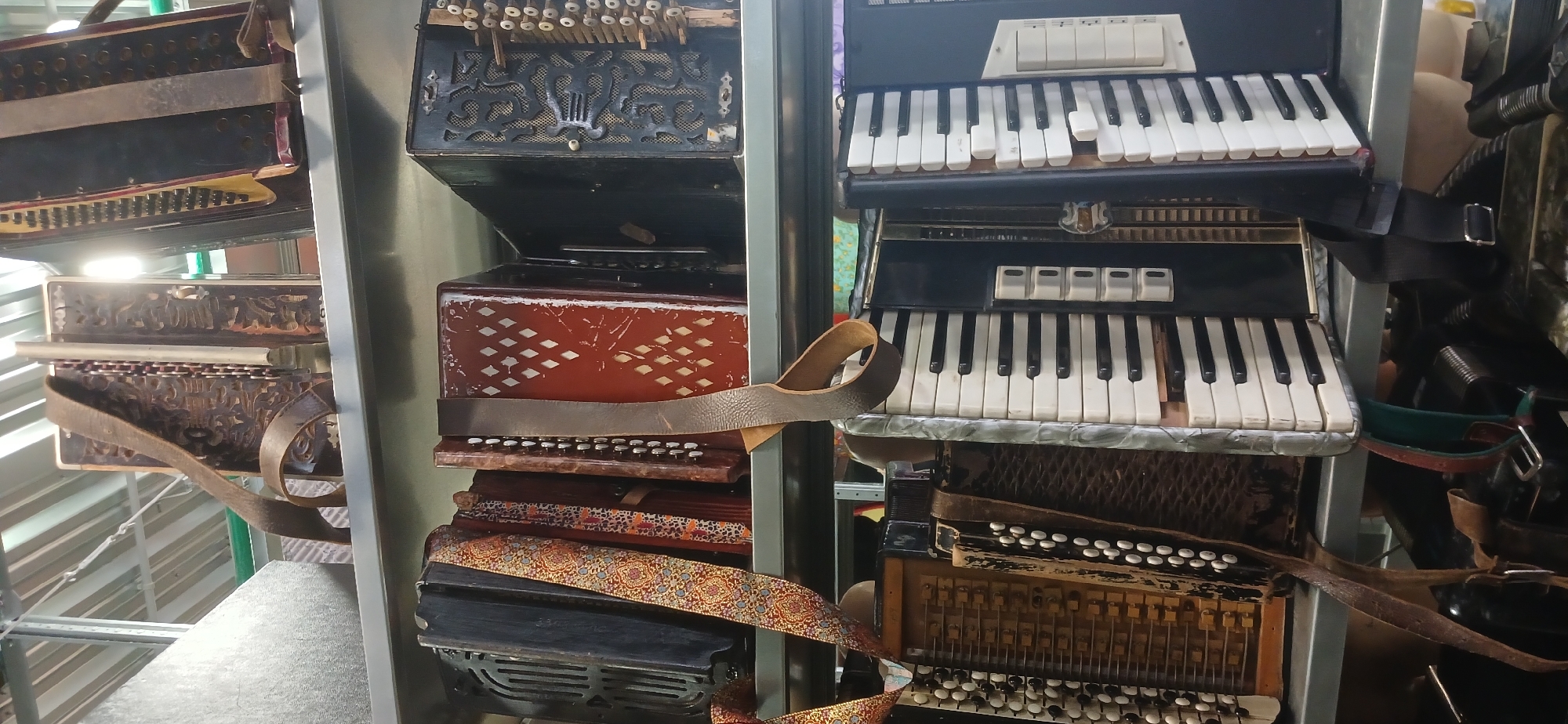 The image shows a collection of accordions and similar instruments displayed on a metal shelving unit. The instruments are of varying styles and conditions, some with visible wear and tear. The setting appears to be indoors, potentially in a music shop or storage area. The presence of diverse accordions suggests a specialized collection or sale of musical instruments. There is no indication of it being located in Moscow, Russia, or any people being present.