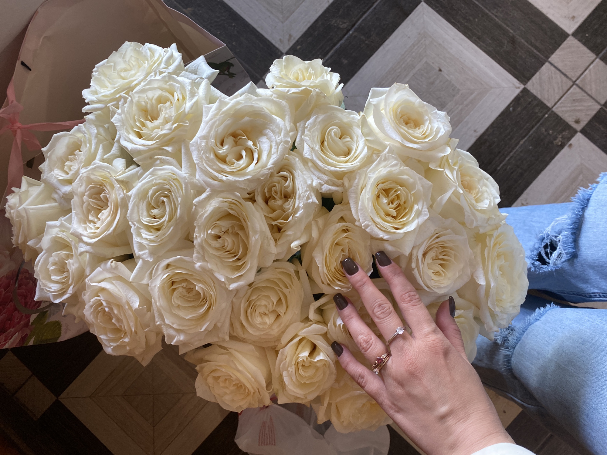 A high-angle shot shows a bouquet of pale yellow roses being held by a person wearing ripped light blue jeans. The hand of the person, adorned with two rings and dark nail polish, is resting on top of the roses. The bouquet is placed on a black and white checkered floor. The background also shows a glimpse of a floral patterned box and a pink ribbon. The scene appears to be indoors, possibly in a home. No specific location in Chaikovsky, Russia, can be determined from the image.